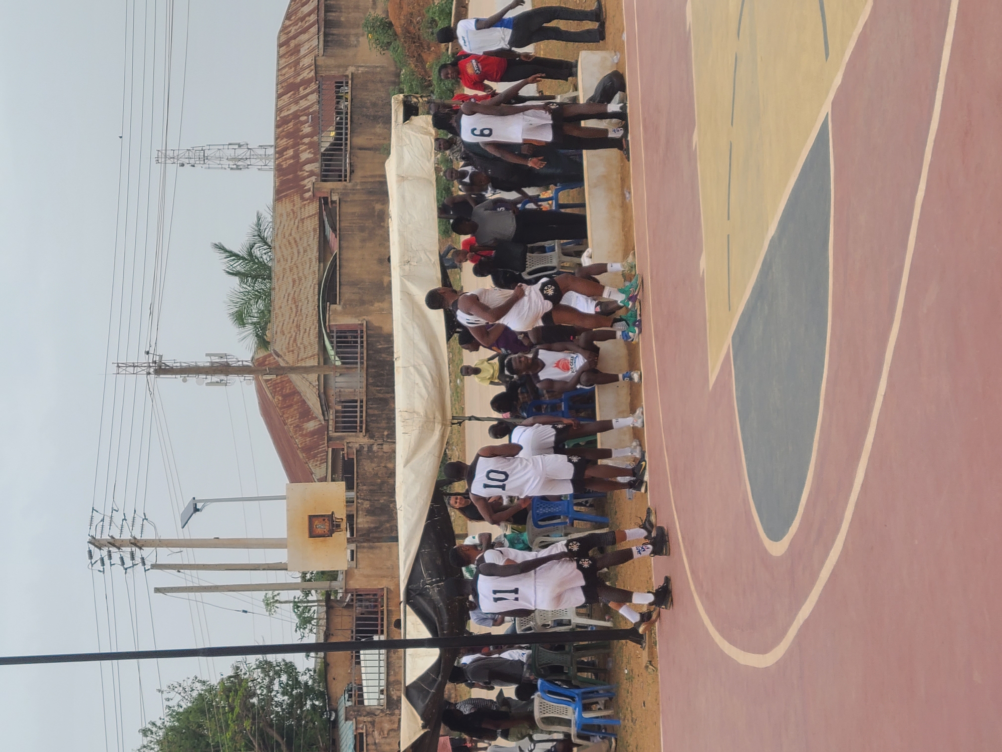 The image shows a basketball court with players seated on chairs along the sidelines. The players are wearing white jerseys with numbers. Behind the players, there is a temporary tent-like structure. Beyond the tent is a weathered building with a corrugated iron roof and multiple windows. Power lines and utility poles are visible in the background. The basketball court has painted sections in red, yellow, and blue. The overall location appears to be an urban area, likely in Jos, Nigeria. The setting is outdoors, seemingly during a game or practice session.