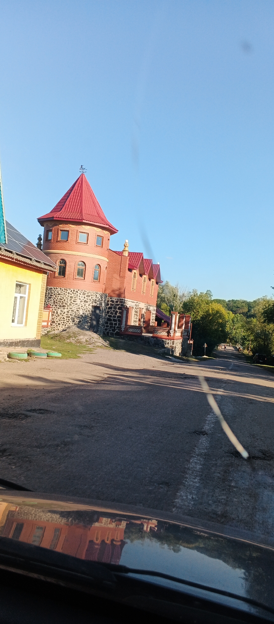 The image depicts a bright, sunny day in Ternivka, Ukraine, focusing on a unique, castle-like building constructed of red brick and stone. The building has a prominent red conical roof topped with a weather vane, multiple arched windows, and a lower section of rough-hewn stone. To the left of the main structure, a yellow building with a section of solar panels on its roof is partially visible. The foreground shows a paved road with patches of damage and an asphalt reflection of the surrounding scenery, including the distinctive building. The background features a line of lush green trees against a clear blue sky. The overall scene suggests a residential or recreational property, possibly a tourist attraction, due to its unconventional architecture.