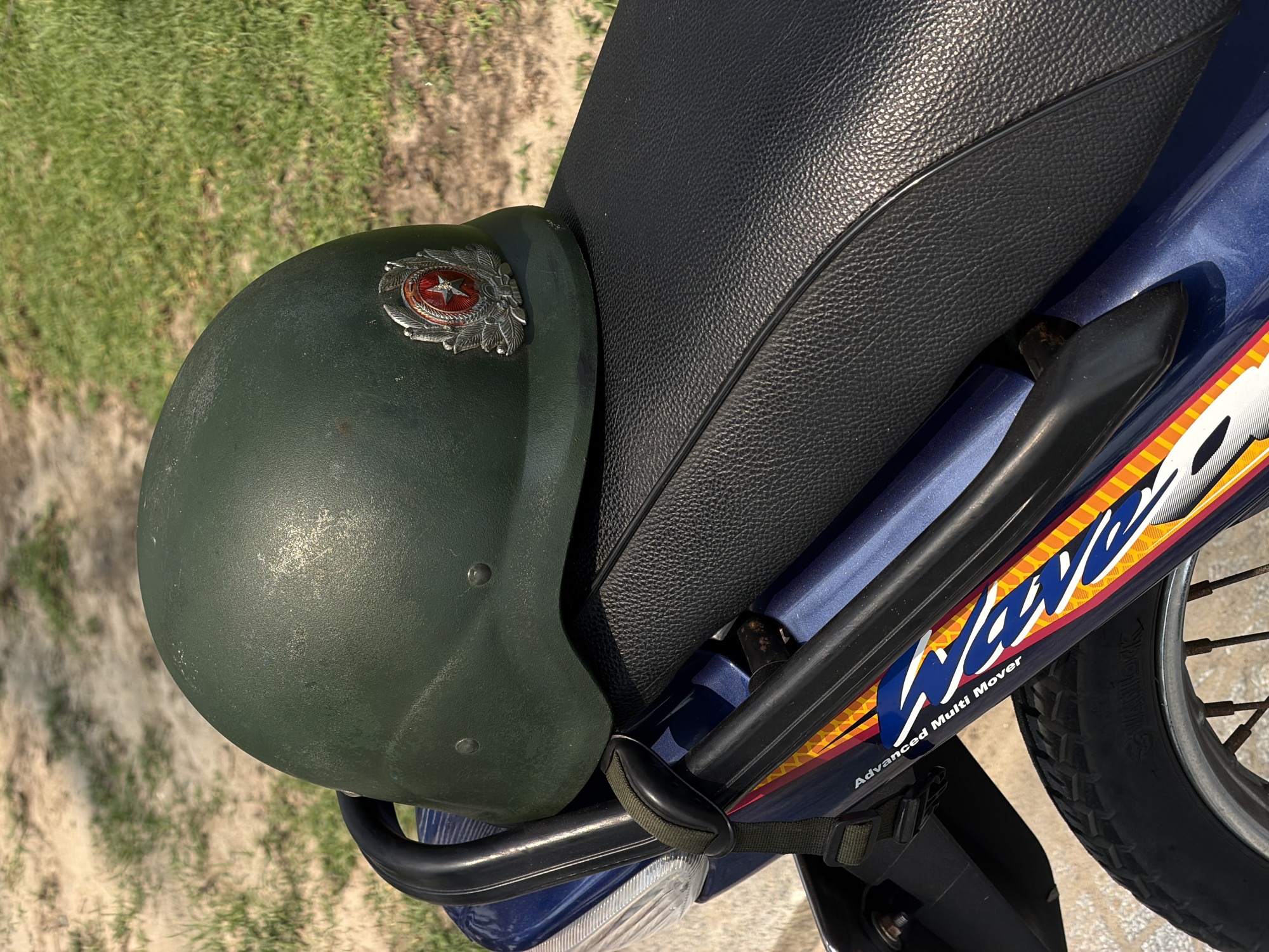 A green military helmet bearing a star emblem rests on the seat of a blue motorcycle in an outdoor setting. The motorcycle has the text "Wave Advanced Multi Mover" printed on its side. The helmet appears slightly worn. The location is unspecified, but the presence of the military helmet and motorcycle suggests a potential location in Vietnam. The image does not contain any visible people or active interactions. The environment consists of a paved or dirt surface surrounded by grassy areas.