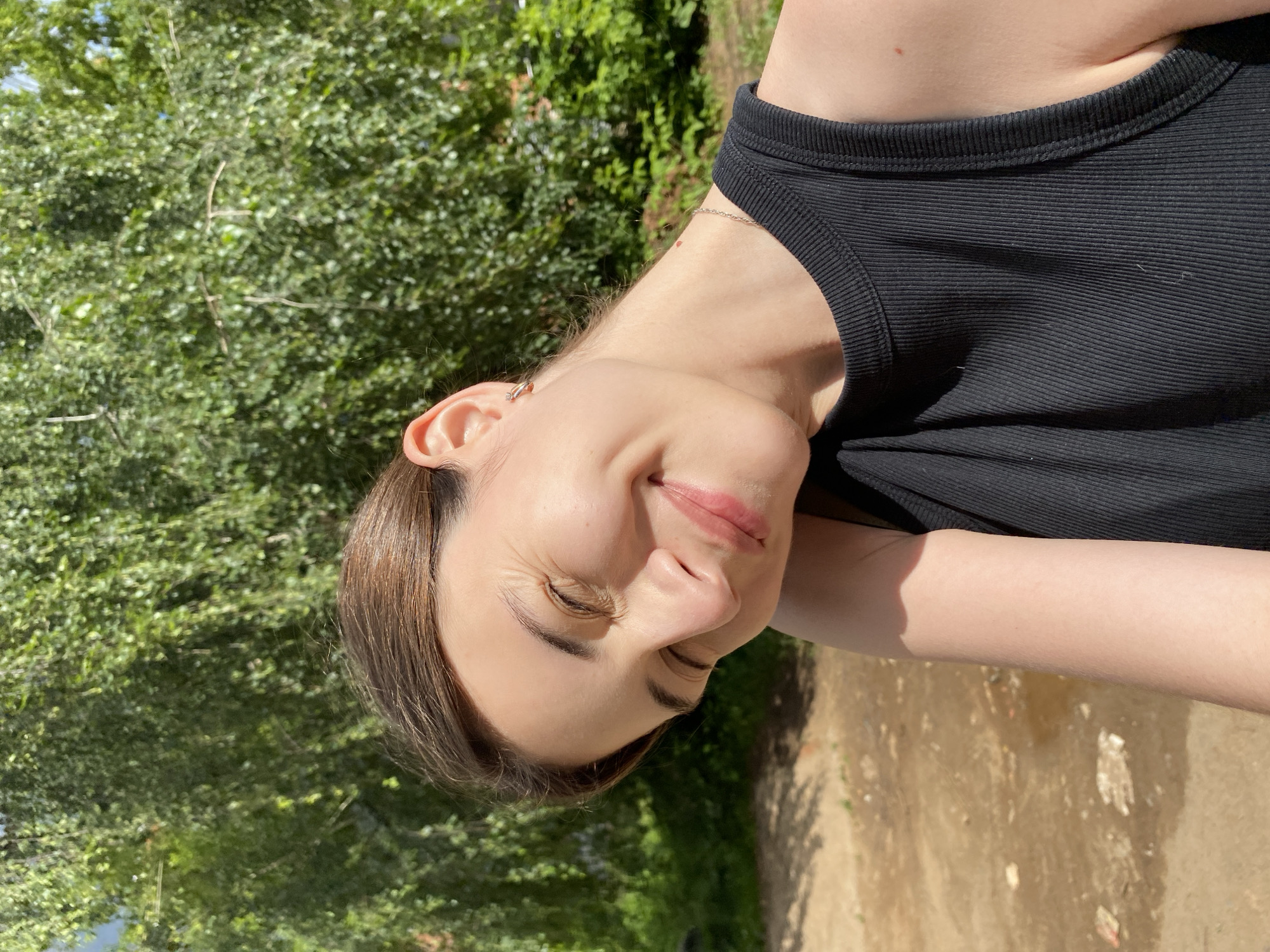 A young woman is captured in a close-up, tilted portrait against a backdrop of lush green foliage and a dirt path. She is wearing a black ribbed tank top. Her eyes are closed, and she has a slight smile on her face, suggesting a moment of contentment or enjoyment in the sun. Her hair is dark brown and pulled back. A delicate chain necklace is visible around her neck, and a small hoop earring adorns her ear. The lighting indicates it is daytime, likely sunny, as evidenced by the bright illumination and shadows. The setting appears to be outdoors, possibly in a park or garden, given the natural surroundings. There is no visible text in the image.