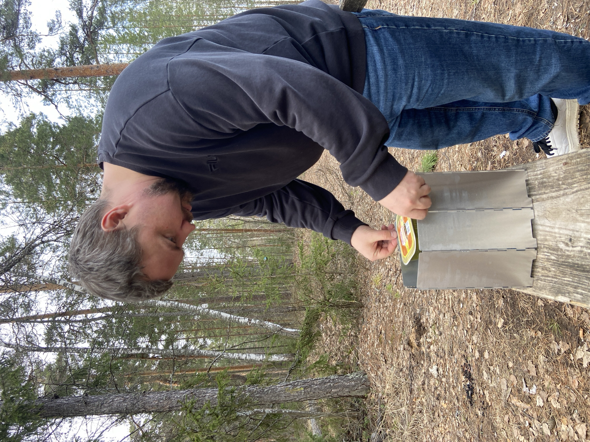 A man in a black sweatshirt and blue jeans is bending over, placing a yellow-labeled jar into a metallic container with multiple sections. The container is sitting on a weathered wooden surface, possibly a bench or table, surrounded by dry leaves and sparse vegetation. The environment appears to be a forest or wooded area, with tall trees visible in the background. The photo does not provide enough context to confirm the location in the city of Chaikovsky, Russia.