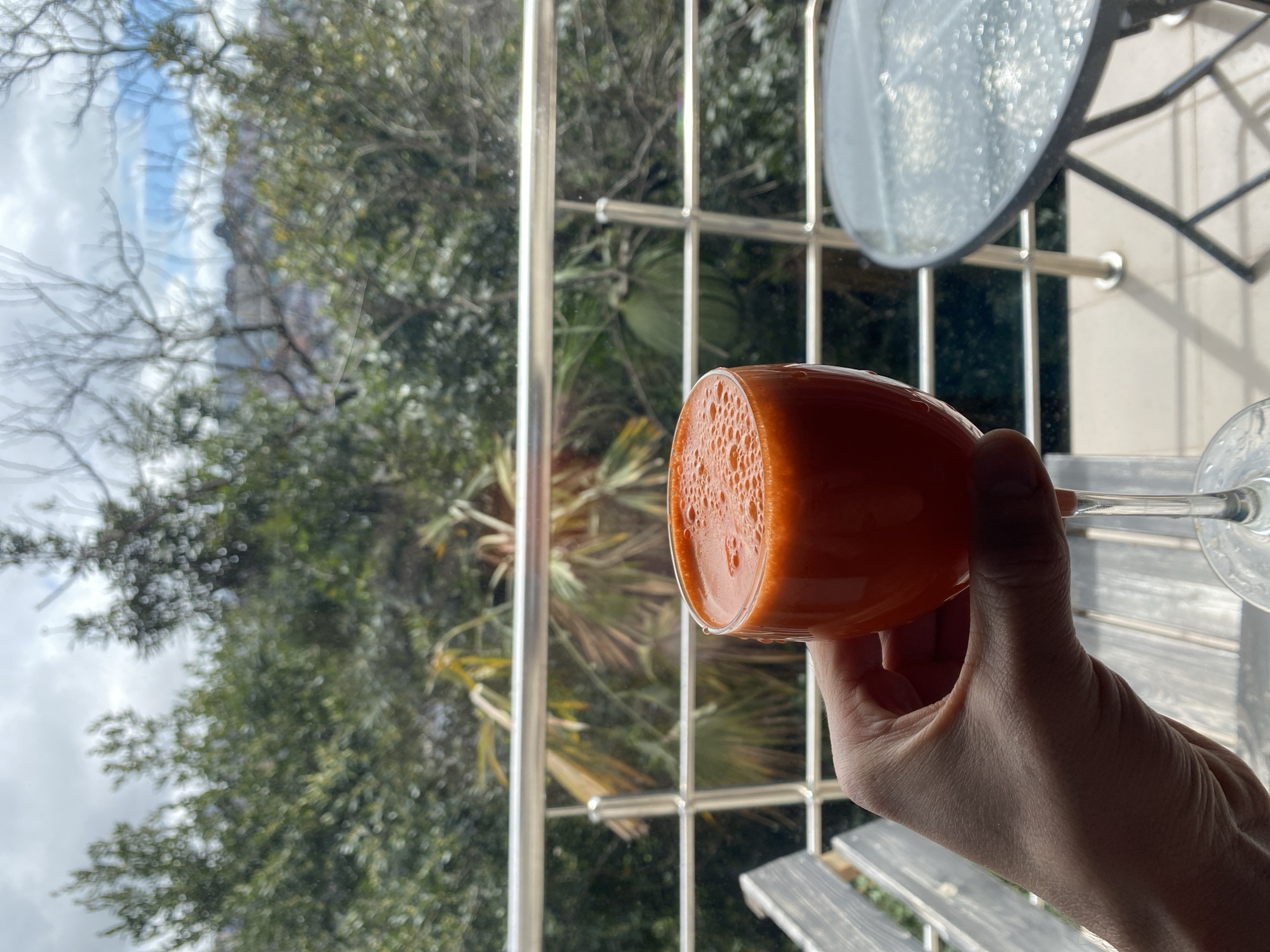 A person's hand holds a glass of orange juice with bubbles in it against a blurred background. The background shows a metal window grill or fence, green trees and a hint of a mountainous landscape with a cloudy sky. The location appears to be a balcony or outdoor area in a location with subtropical or Mediterranean vegetation. Based on the hints, this is taken in Sochi, Russia. A round, reflective surface (possibly a table top) is partially visible at the upper right. The surface beneath the glass appears to be a wooden slatted table.