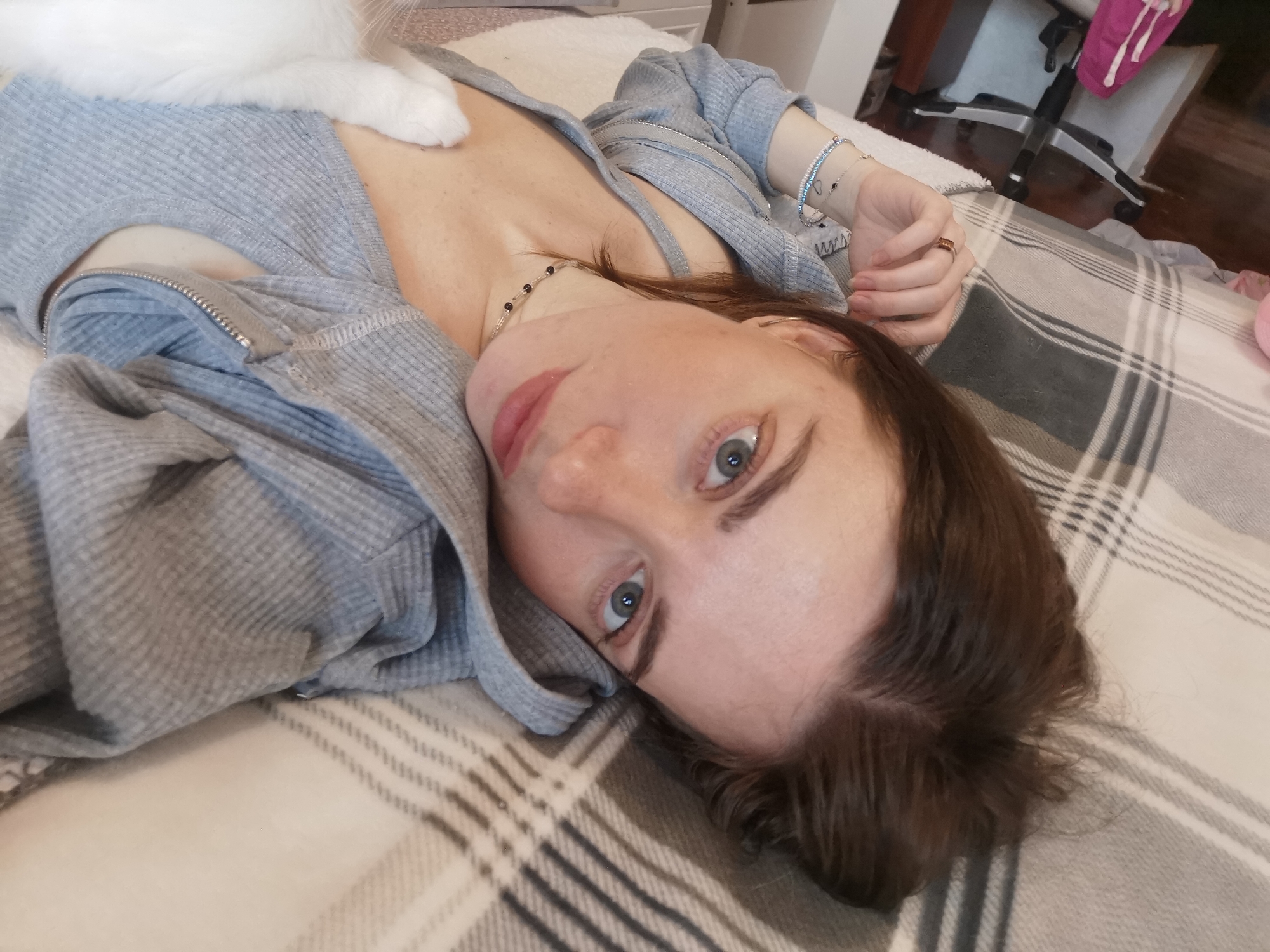 A woman is lying on a plaid blanket, wearing a gray zippered top, with a white cat paw resting on her chest. She is looking directly at the camera. The background includes a chair and flooring. The location is an indoor setting.
