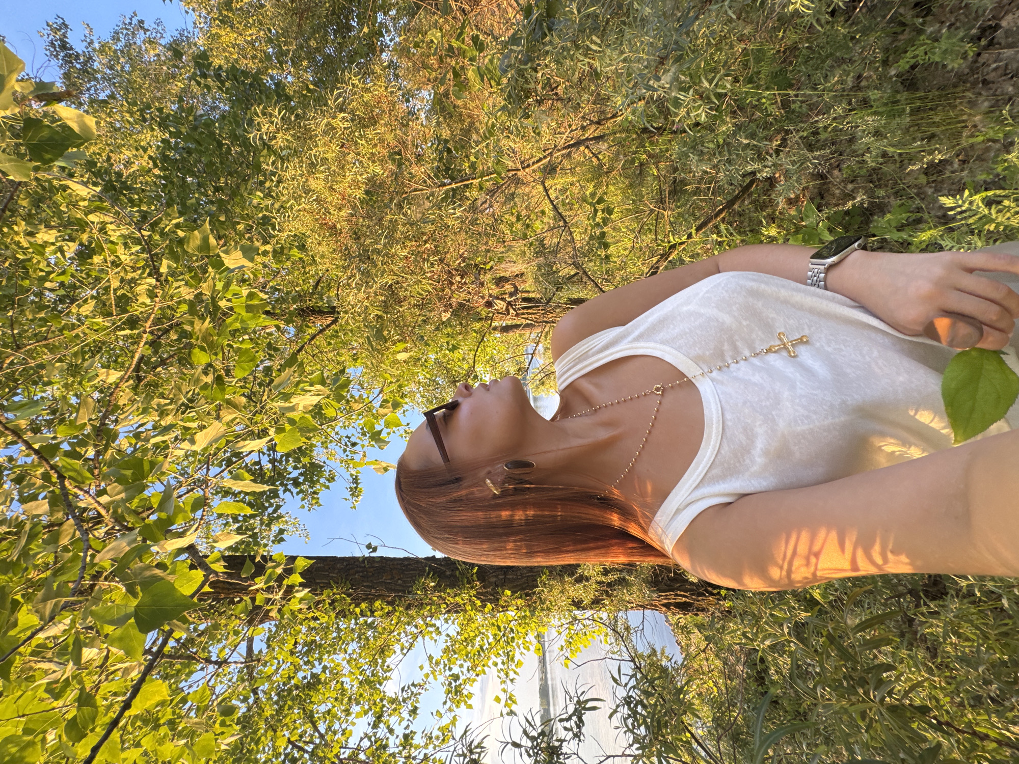 A woman with long, auburn hair and sunglasses stands outdoors, facing to her right and looking up. She is wearing a white tank top, a gold necklace with a cross pendant, a large ring on her right hand, and a wristwatch. The woman is positioned in front of a tree with dappled sunlight on her arm. Behind her, to her left, is a body of water. The background is filled with lush green foliage. The scene appears to be in a natural setting, possibly a park or a natural area, with the water and trees suggesting a lakeside or riverside location. The lighting indicates it is likely late afternoon or early evening. There is no visual information to suggest this location is in Noviy, Russia.