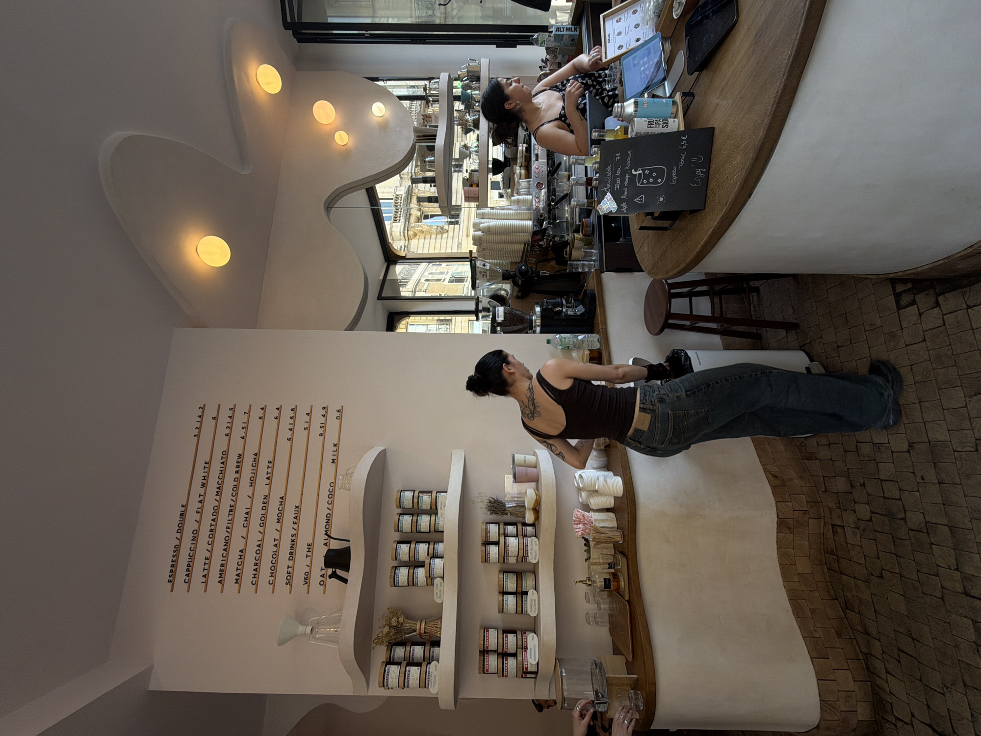 The image depicts the interior of a modern cafe with a minimalist aesthetic. Two women are visible behind a wooden counter. One woman, wearing a dark tank top and jeans, stands with her back to the camera, facing the counter. The other woman, wearing a patterned dress, is seen from the side, also behind the counter, engaged with the space.
The cafe features white, wavy architectural elements on the ceiling and walls, punctuated by circular yellow light fixtures. A menu board with a list of coffee and drink options is mounted on the white wall. Shelves above the counter display various jars and containers, suggesting retail of cafe products. On the counter, there are coffee machines, cups, straws, bottles, and a tablet.
The flooring is made of patterned wooden tiles. In the foreground, a portion of the counter curves towards the viewer, displaying a small chalkboard sign with handwritten menu items and prices. The overall atmosphere is clean and contemporary, with natural light filtering in from a large window or doorway in the background, revealing a glimpse of an urban streetscape with buildings.