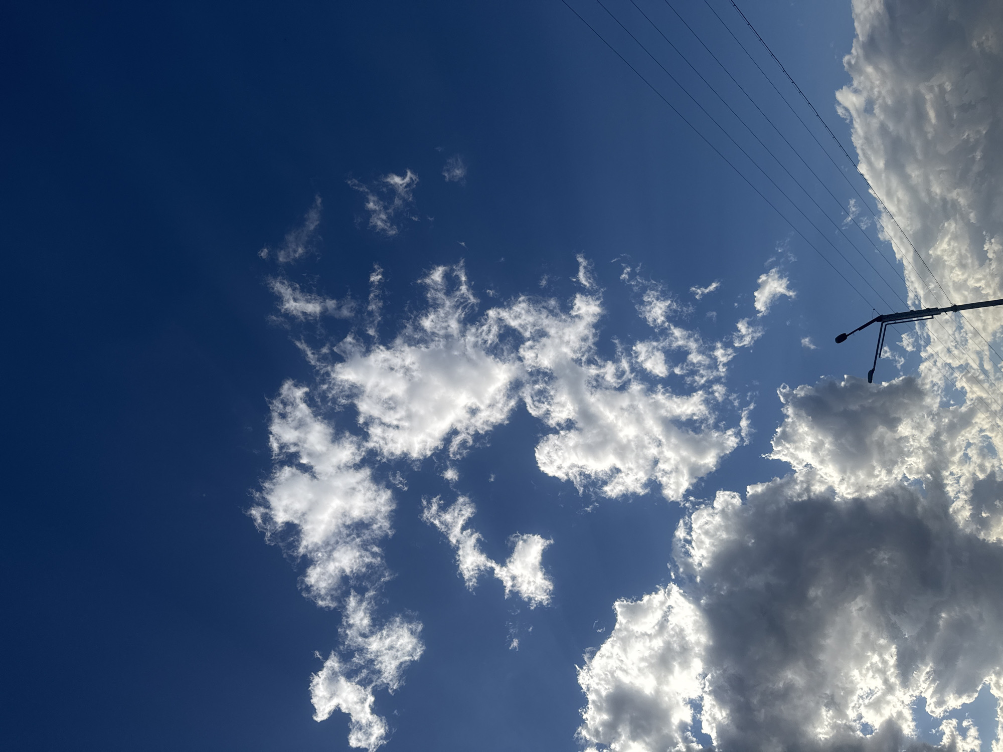 The image captures a view of the sky in Tbilisi, Georgia. A deep blue sky is partially covered with white and gray clouds. Electrical power lines and a street light extend into the scene from the upper right corner.