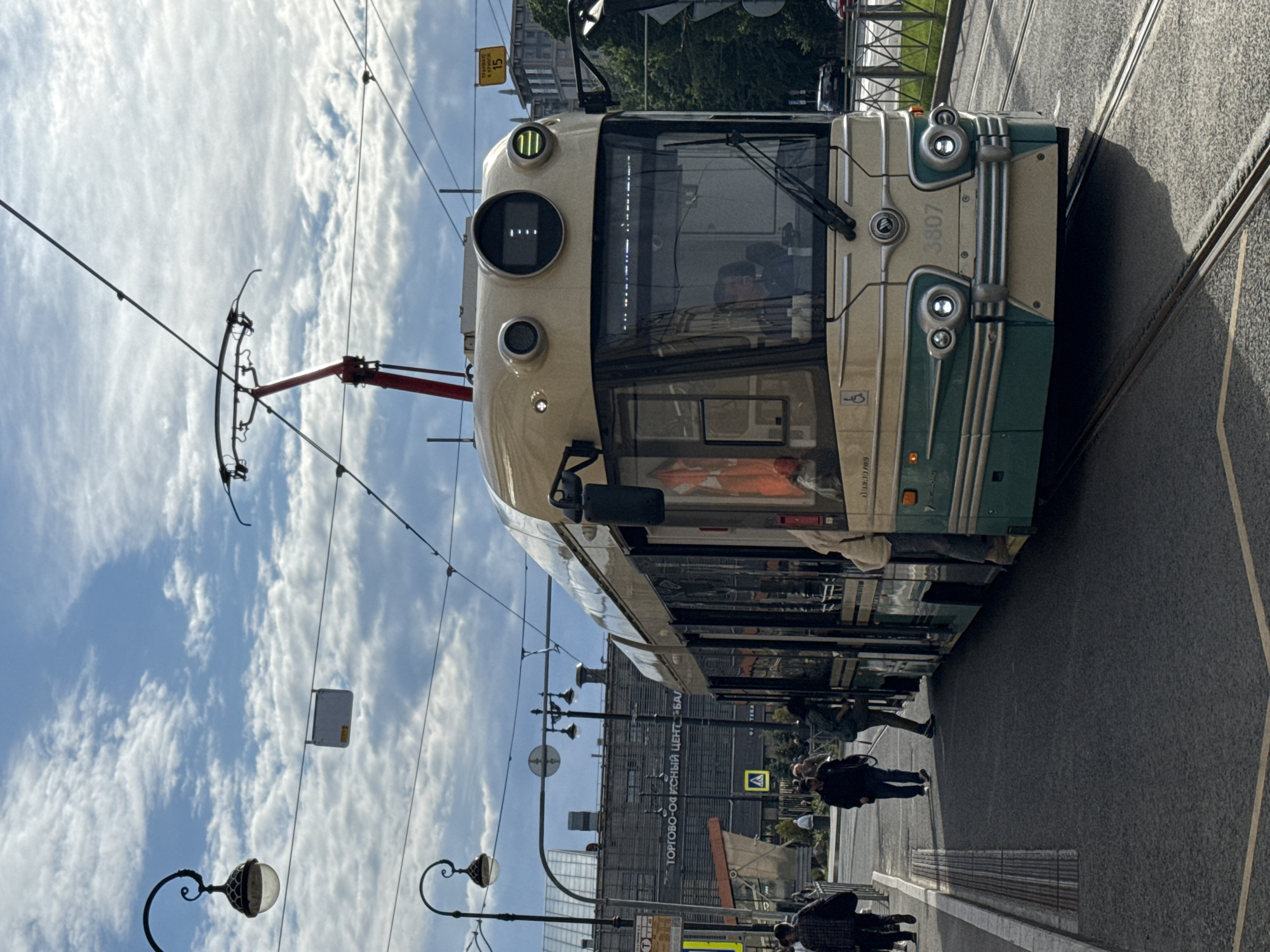 The image depicts a street scene in Saint Petersburg, Russia, likely on a cloudy day. A beige and green tram, numbered 3807, is the central subject, positioned on tram tracks and appearing to be stopped. The tram has a distinctive retro design with circular headlights and a prominent front panel. A driver is visible inside the tram's cabin.
Several people are present on the sidewalk to the left of the tram, some walking and others appearing to be boarding or disembarking. One individual is wearing an orange safety vest. The background reveals a large, dark building with Cyrillic text that translates to "TRADE-OFFICE CENTER 'BAL'." Streetlights with ornate designs are visible along the sidewalk, and overhead power lines for the tram crisscross the sky. A yellow pedestrian crossing sign with an exclamation mark is also visible. The general environment suggests an urban setting with public transportation infrastructure.