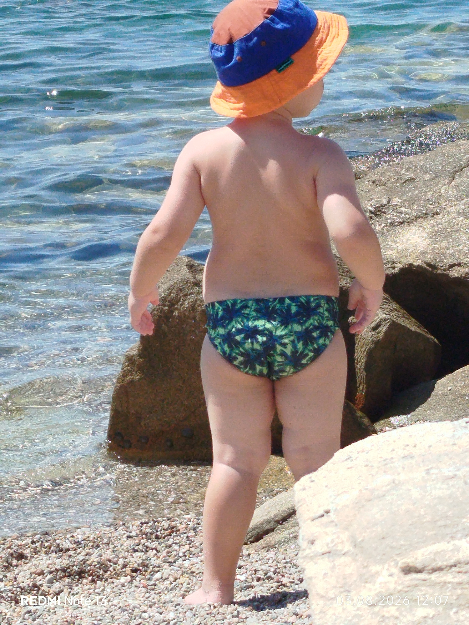 A young child, seen from the back, stands on a pebbly beach near the clear, shallow water of Sithonia Municipal Unit, Greece. The child wears a colorful bucket hat with orange and blue sections, and green patterned swim trunks. The child's skin is smooth and unblemished. They stand next to several large, rough rocks and appear to be looking out at the water. The water is calm with ripples, and the seabed is visible through the clear surface. The environment is sunny and appears to be a warm day. The image captures a moment of quiet observation by the child at the edge of the sea.