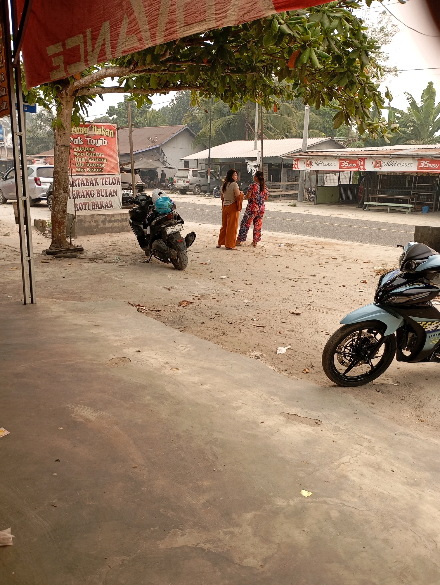 In Sigi, Indonesia, a street scene unfolds under a covered awning with a red and white striped banner that reads "JIANCE". Two women stand on a sandy area near a tree, one wearing an orange outfit, the other a patterned dress. In the background, a few buildings with corrugated roofs line the street, with a sign that reads "PAK TOYIB". Several parked motorcycles are visible, including a black scooter with two helmets and a light blue Yamaha motorcycle in the foreground. The ground is a mix of concrete and sandy dirt, with scattered debris.