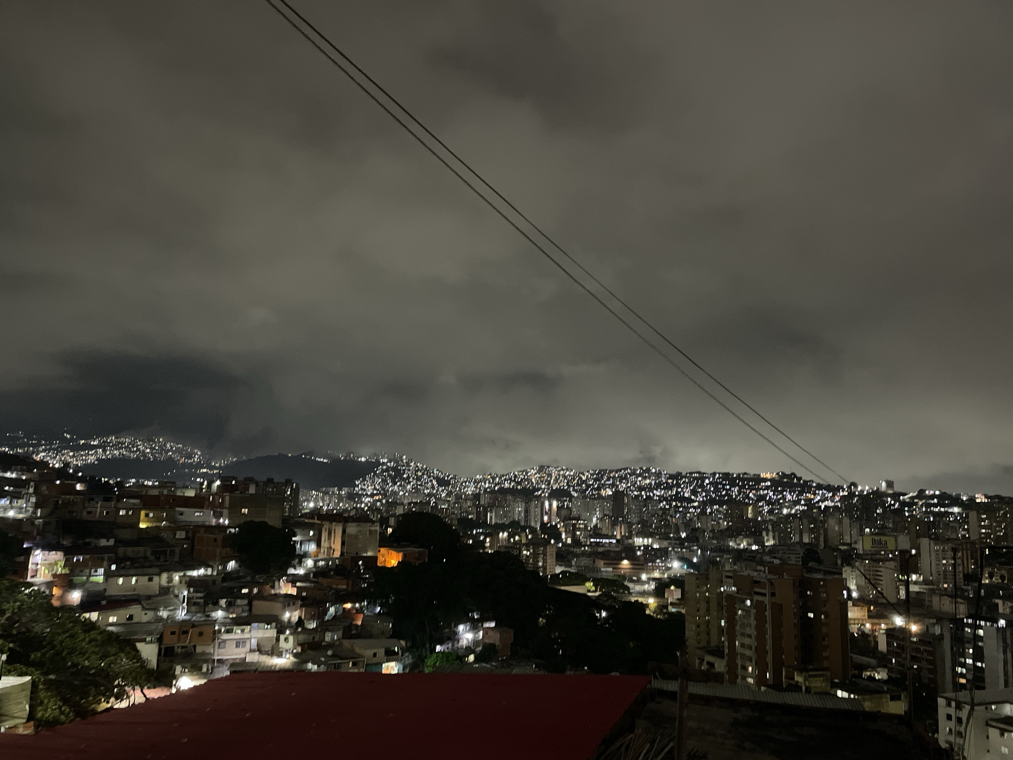 The image depicts an urban landscape at night, likely in Caracas, Venezuela, given the city's characteristic topography and density. A red roof is visible in the foreground. The middle ground shows a densely populated area with a mix of low-rise buildings and taller apartment blocks, illuminated by scattered lights. In the background, hills are covered with more dense housing. The sky is overcast with heavy clouds, and two power lines traverse the upper portion of the image.