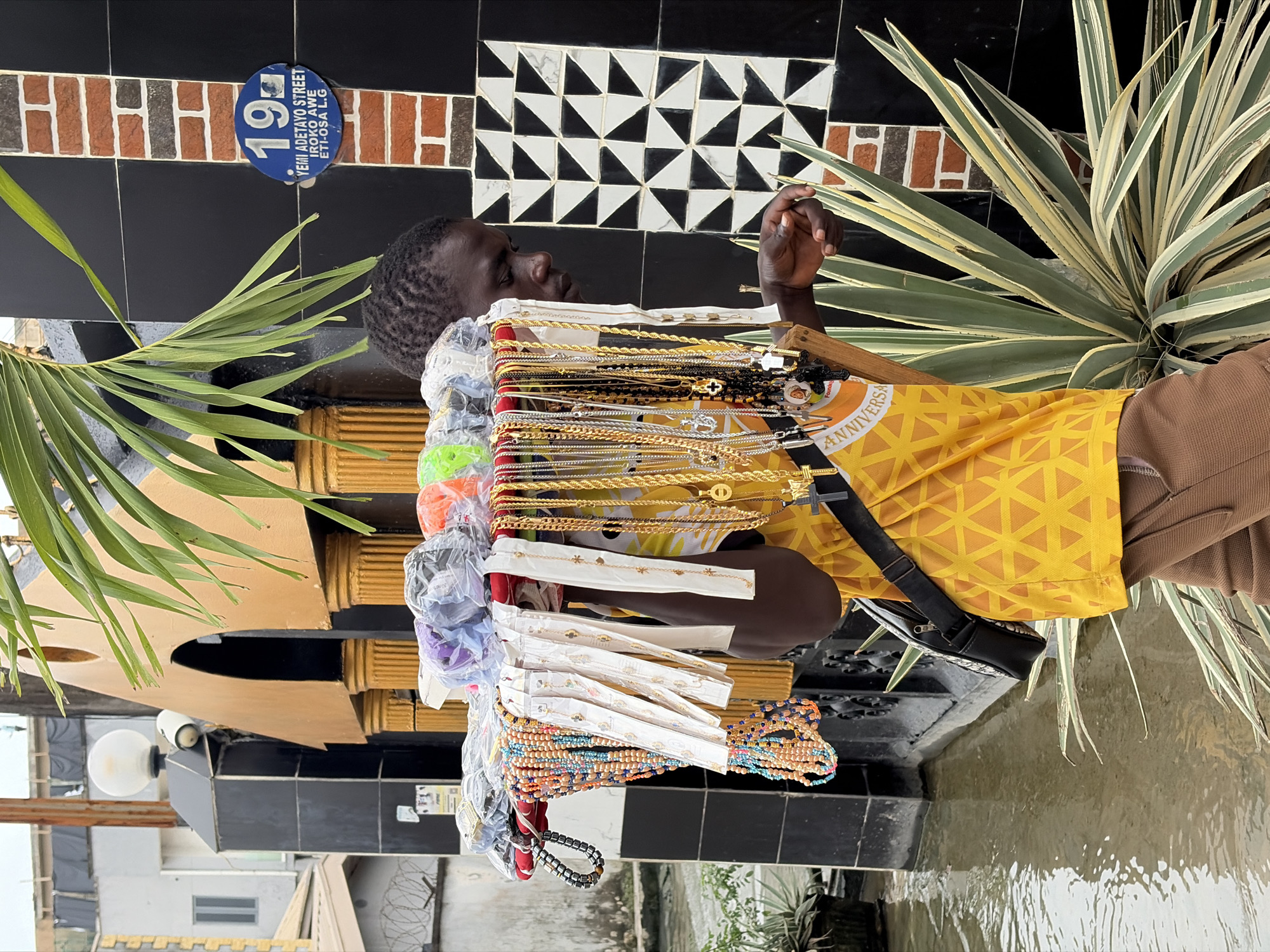 In Igboefon, Nigeria, a person is displaying a selection of necklaces and bracelets. The display includes numerous gold-colored chains of varying thickness, some with pendants such as crosses. There are also bracelets and necklaces made of colorful beads. The items are arranged on a stand that appears to be made of wood. The person, wearing a yellow shirt with a geometric pattern and brown pants, is reaching towards the display, possibly to adjust or show an item.
The background features a wall decorated with a mosaic of black and white triangles, interspersed with bricks. A blue oval sign with white text is mounted on the wall, reading "19 YEMI ADETAYO STREET EROKO AWE ETI-OSA L.G". Adjacent to the wall is a lush green plant with long, striped leaves.
The scene appears to be outdoors, with evidence of water on the ground, suggesting it might have rained recently or is a wet environment. The lighting suggests it is daytime. The overall atmosphere is that of a street vendor or market stall. The person's posture and focus suggest they are engaged in their business, presenting their wares.