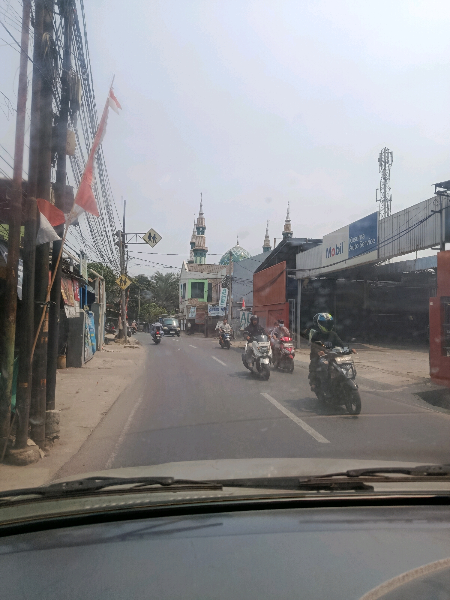 This image depicts a street scene in East Jakarta, Indonesia. In the foreground, the dashboard and windshield of a vehicle are visible, indicating the image was taken from inside a car. The road in front is paved and shows white lane markings. Several motorcycles are in motion on the road, some carrying passengers.
To the left of the road, there is a sidewalk and a row of buildings, including a structure with vertical wooden poles and a red and white Indonesian flag draped from one of them. Further down the street, a mosque with a green dome and minarets is prominent. There are also utility poles with numerous wires.
On the right side of the road, a building with a large blue sign reading "Mobil KUSUMA AUTO SERVICE" is visible. Further down, a cell tower stands against the hazy sky. The overall environment suggests a typical urban street in Indonesia, characterized by a mix of residential, commercial, and religious structures, with a high volume of motorcycle traffic. The weather appears to be overcast or hazy.