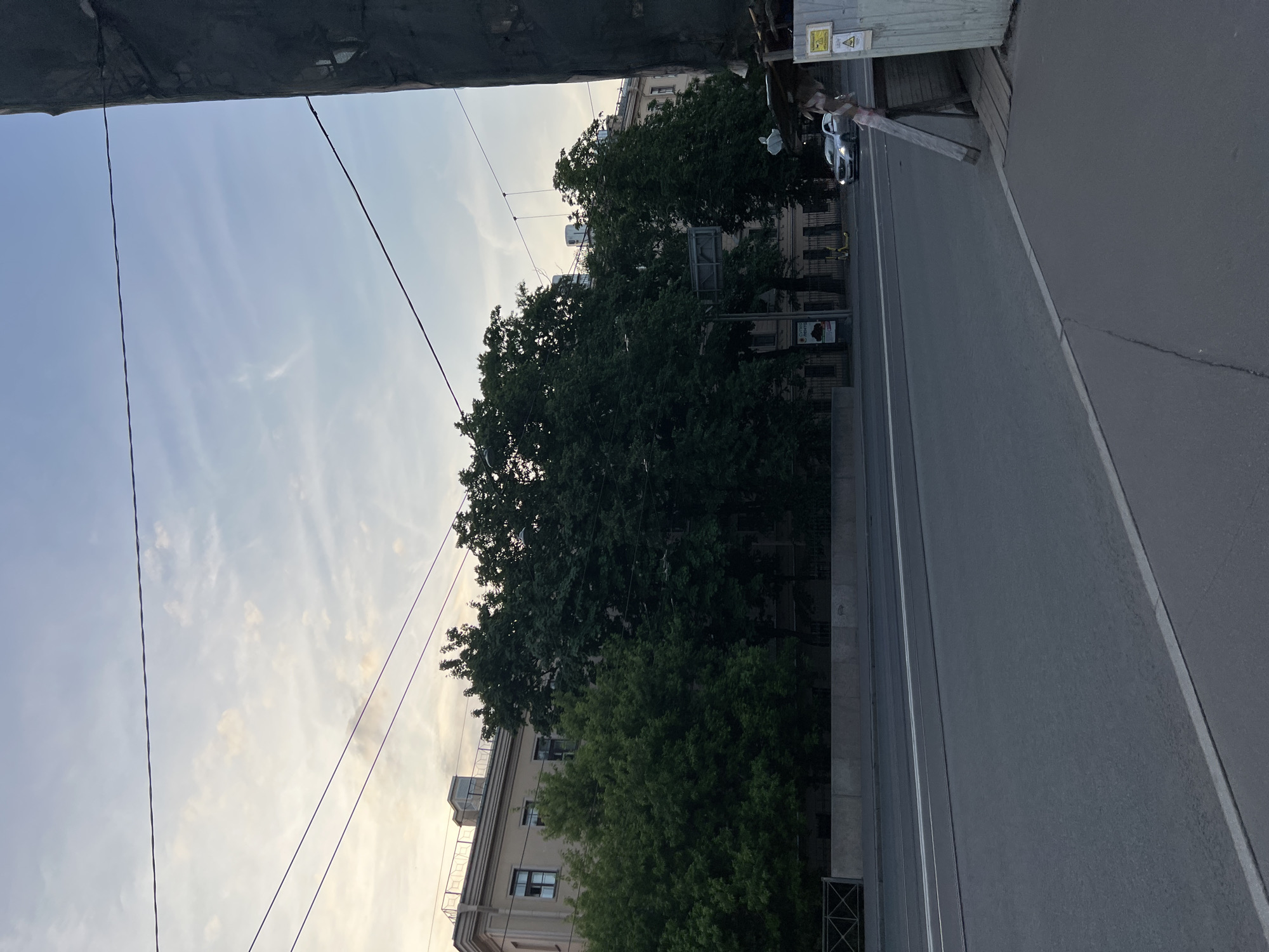 The image captures an urban street scene in an unknown city in Russia. The foreground features a paved road with white lane markings and a pedestrian sidewalk. To the left, a concrete barrier separates the road from a grassy area with trees and a fence. Behind the trees, several buildings are visible, characterized by their light-colored facades and a rooftop structure with railings. The middle ground shows a row of dense green trees lining the street. A silver car is driving on the road. In the background, the sky is partly cloudy with the soft hues of dusk or dawn. On the right side of the image, a temporary structure made of wood and metal sheeting is partially visible, with warning signs attached to it. Overhead, power lines are strung across the scene.