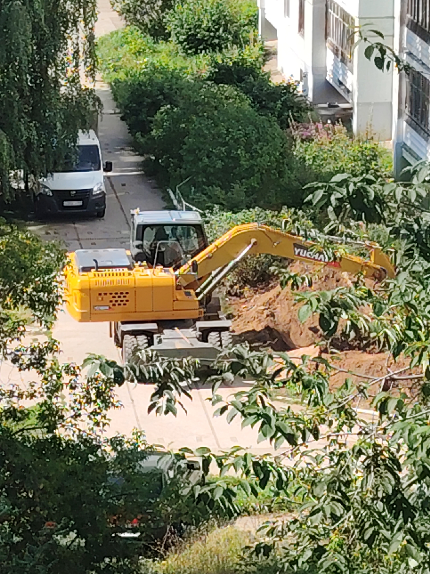 An aerial, slightly blurred view of a construction site in Chaikovsky, Russia, shows a yellow excavator with the brand "YUANHAI" on its arm, actively digging a trench. The excavator is positioned on a dirt pile. To the left, a white van is parked on an asphalt surface. Surrounding the scene are lush green trees and foliage, with a white building visible in the background.