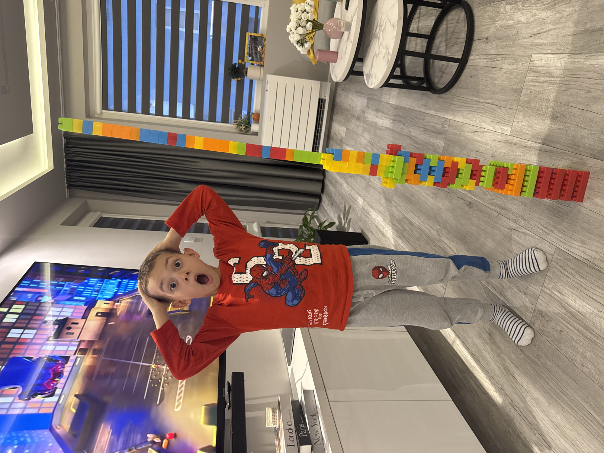 A young boy in a red Spider-Man shirt and gray striped socks stands in a room with light gray, wood-look flooring. He has his hands on his head and is making an surprised facial expression. He's standing near a long line of connected colorful building blocks that stretch from the window with gray blinds to beyond the boy. Behind him is a white cabinet, and on the left, a large television displays an animated scene. In the background, there's a white radiator, a plant, and two small round tables. The location is not discernible from the image.