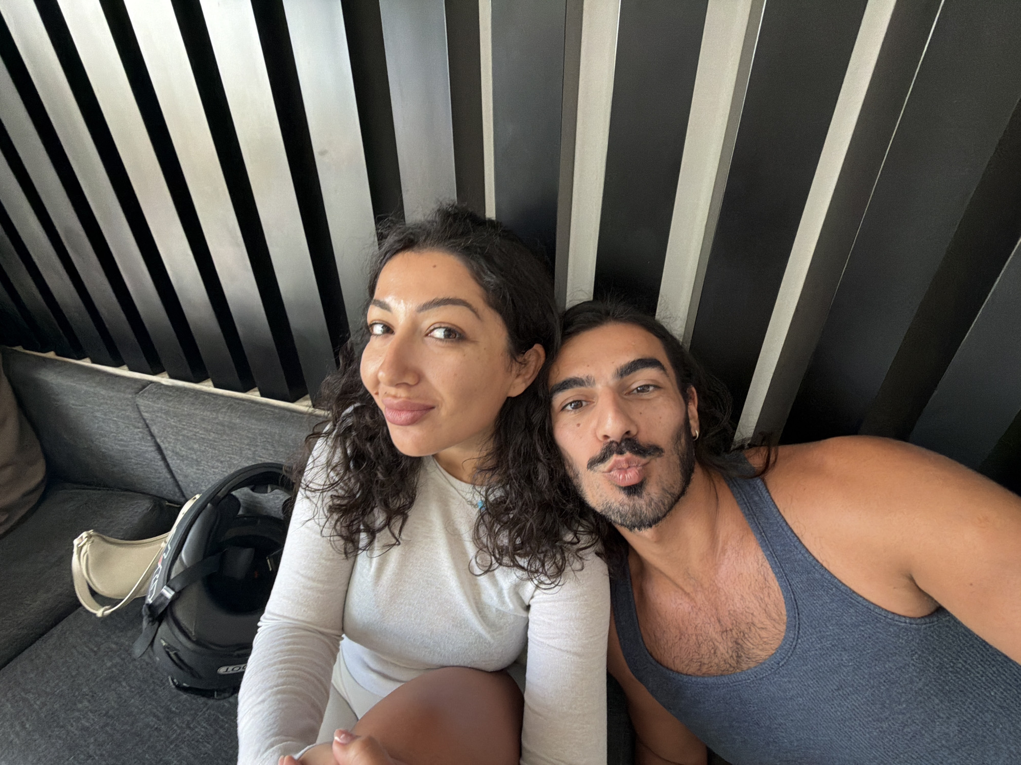 A man and a woman are taking a selfie on a grey couch in front of a wall with black and silver vertical slats. The woman, on the left, has dark curly hair and is wearing a light grey long-sleeved shirt and white shorts. She has her right hand on her lap. The man, on the right, has dark hair and a mustache and beard. He is wearing a grey tank top and is puckering his lips. His right arm is around the woman. On the couch next to the woman is a black motorcycle helmet and a small cream-colored purse. The lighting suggests it is daytime. The environment is an indoor setting, possibly a hotel room or lounge, in Mengwi, Indonesia.