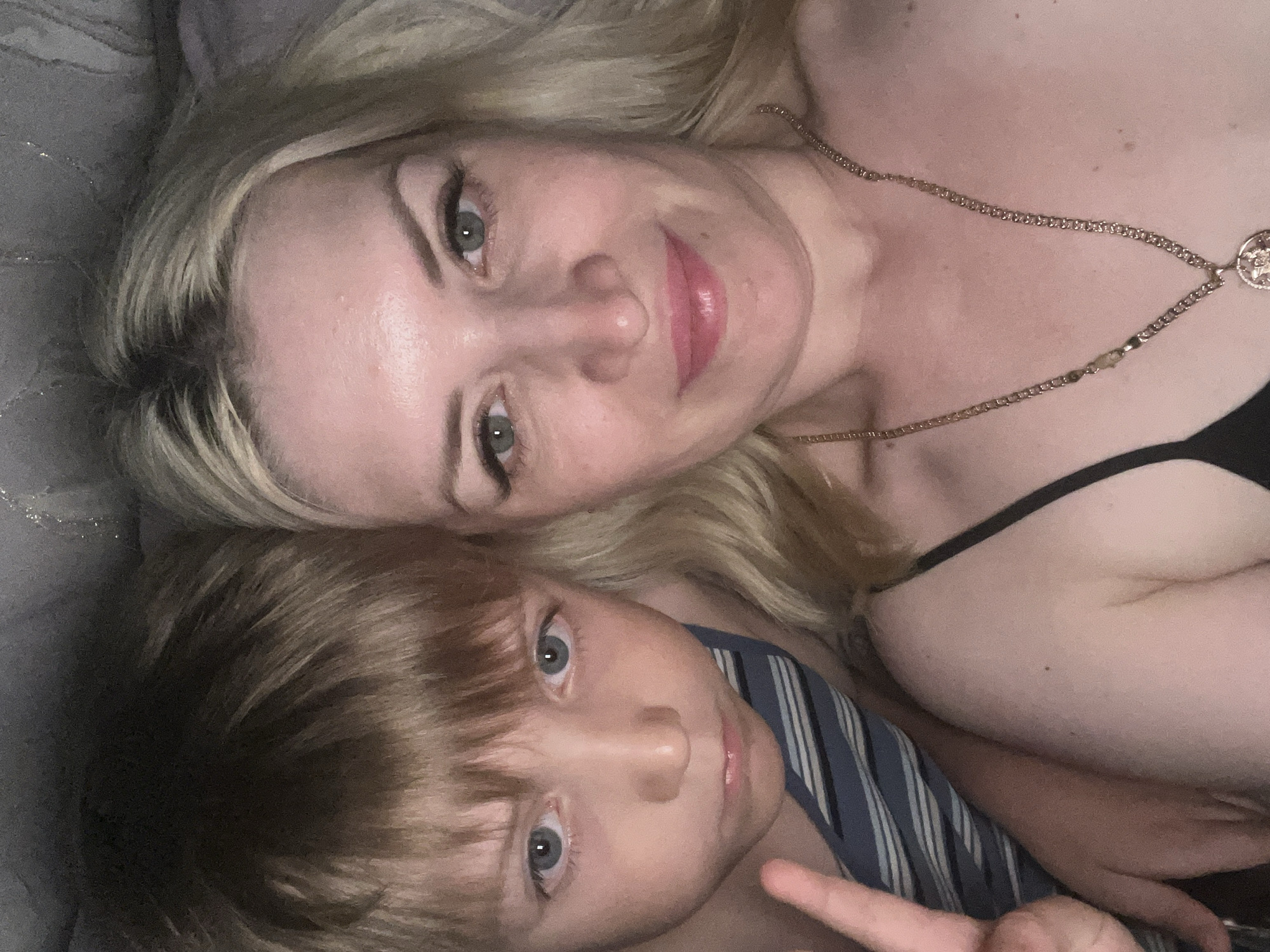 This media file shows a selfie of a woman and a young child, likely mother and son, lying down together. The subjects are close to the camera, with the woman's head and upper body filling the top half of the frame and the child's head in the bottom left. The woman has blonde hair and light-colored eyes. She is wearing a gold necklace. The child has light brown hair and blue eyes. He is wearing a blue and white striped garment. The background appears to be a soft, gray surface, possibly a bed or sofa. The lighting suggests an indoor setting, possibly during the day given the natural light implied, or a well-lit room in the evening. The overall mood appears relaxed and affectionate, with the two subjects posing for a photograph. There is no visible text in the image. The location context is Tyumen, Russia.