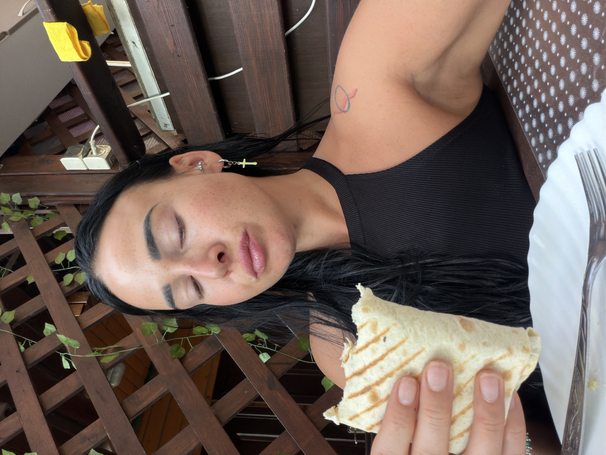 The image features a close-up shot of a young woman with dark hair and closed eyes, holding a partially eaten wrap in her right hand. She is wearing a black sleeveless top and has earrings in her left ear, one of which is a small cross. A tattoo of a blue circle with a red line through it is visible on her upper arm. The background consists of a wooden slatted structure, possibly a fence or pergola, with some green artificial leaves attached. A white tablecloth with a repetitive circular pattern and a fork are also visible on the right side of the frame. The lighting suggests it is daytime, and the overall setting appears to be outdoors, possibly at a cafe or restaurant. The woman's facial expression, with her eyes closed and lips slightly parted, conveys a sense of relaxation or enjoyment.
