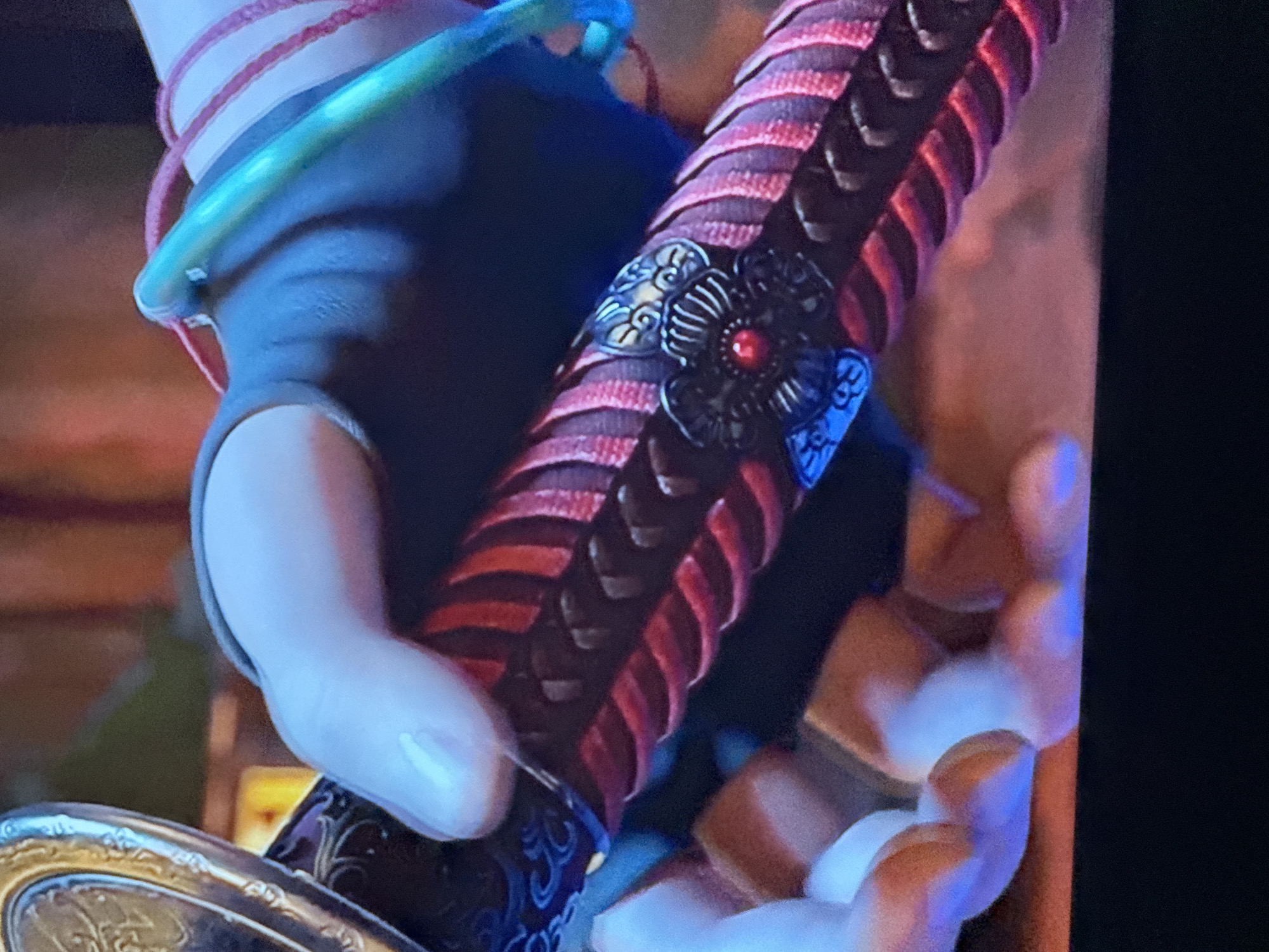 A close-up image shows a hand gripping the handle of a sword, possibly a katana. The handle is elaborately decorated with red and black woven wrapping and ornate metal embellishments, including floral designs and a red jewel. The hand appears to be gloved, with exposed fingers. A metallic, ornate ring is partially visible at the bottom of the handle. There is no evidence indicating the location is in Mytishchi, Russia.