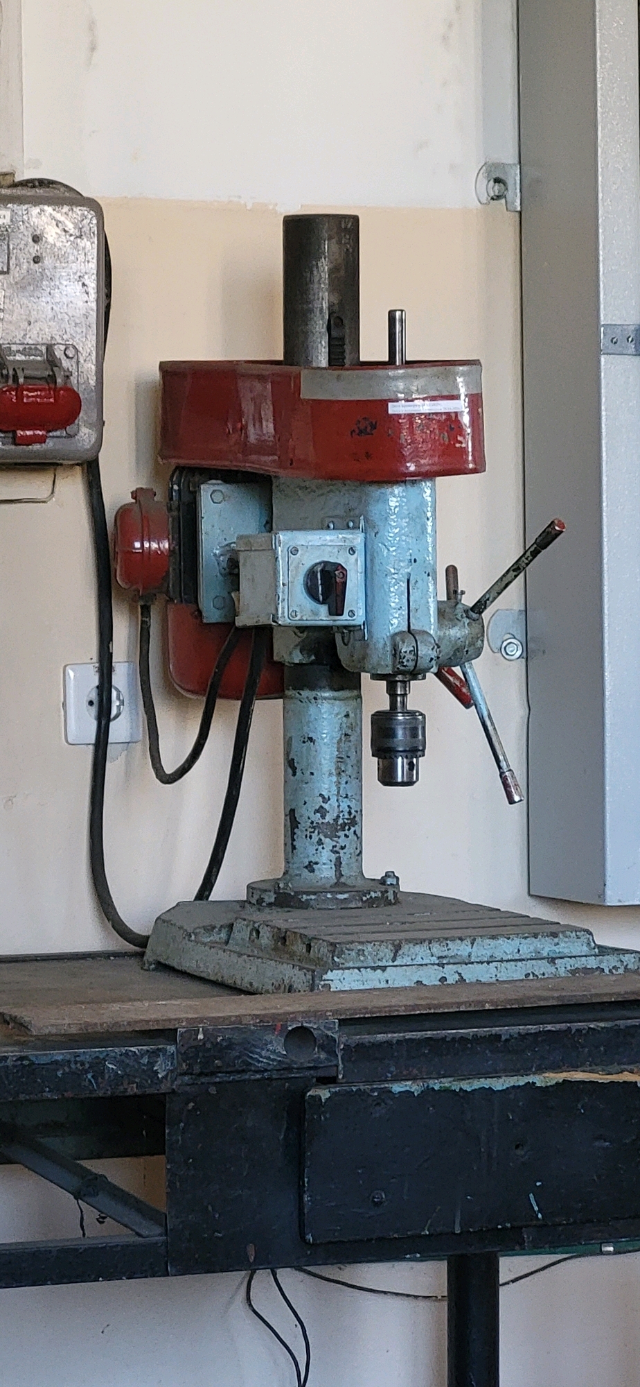 An industrial drill press, painted light blue with a red shroud and a weathered gray base, is situated on a dark wooden surface. The drill press features a chuck, a height adjustment lever, and a power switch. To the left of the drill press, a silver metal electrical box with red buttons and a coiled black power cord is mounted on a light-colored wall. The wall also has a white electrical outlet. The drill press and surrounding area appear to be in an indoor workshop or industrial setting. The image does not provide any specific information about its location in Oskemen, Kazakhstan.