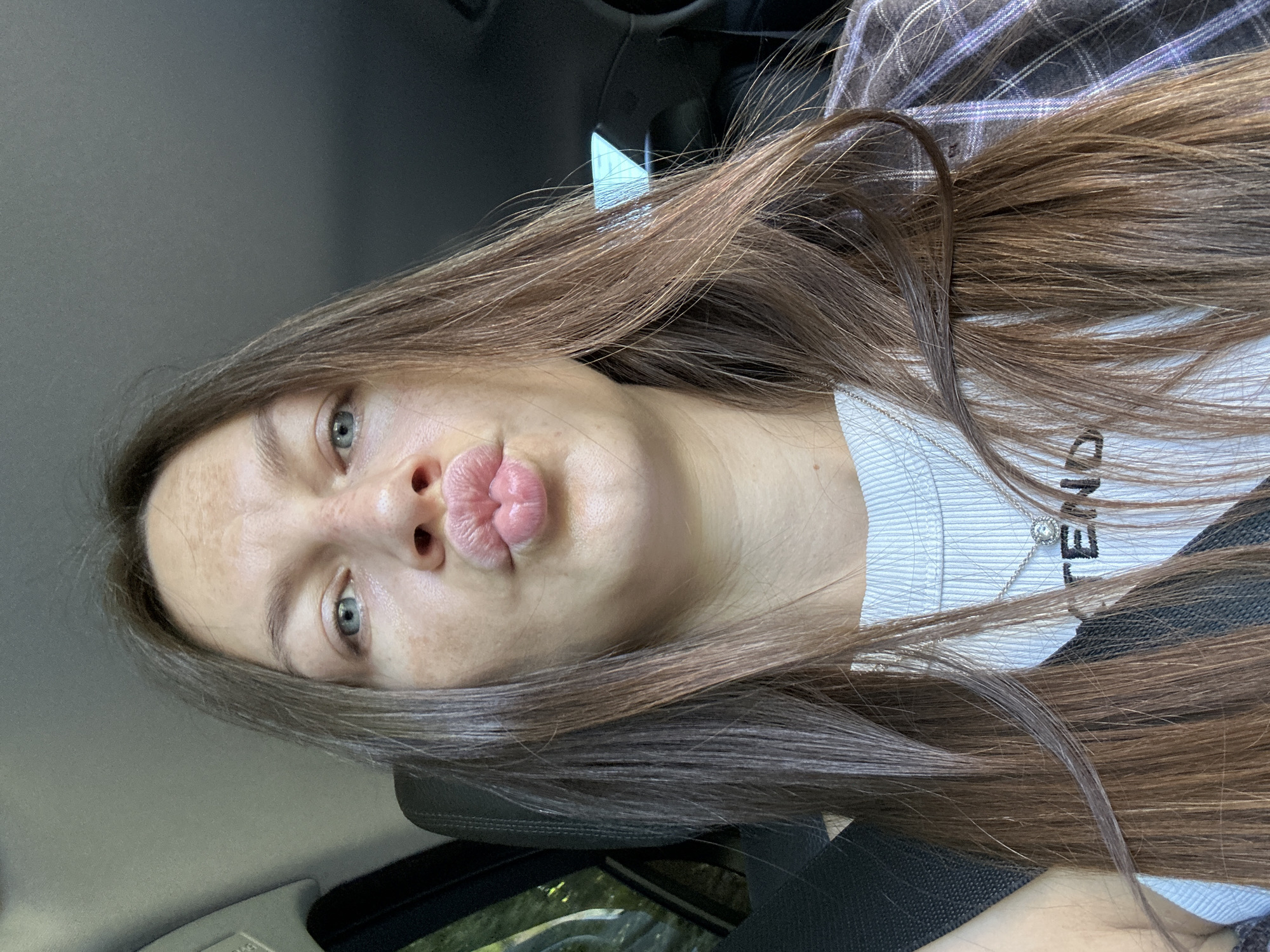 A young woman with long, brown hair and light blue eyes, wearing a white ribbed tank top and a plaid shirt, is sitting in the driver's seat of a car. She is making a kissing face, with her lips pursed. The interior of the car is visible, including the seatbelt and part of the dashboard. Outside the car window, greenery can be seen, suggesting a natural or semi-rural setting. The image appears to have been taken during the daytime. The specific location of Jabuka, Montenegro is not discernible from the visual information provided in the image.