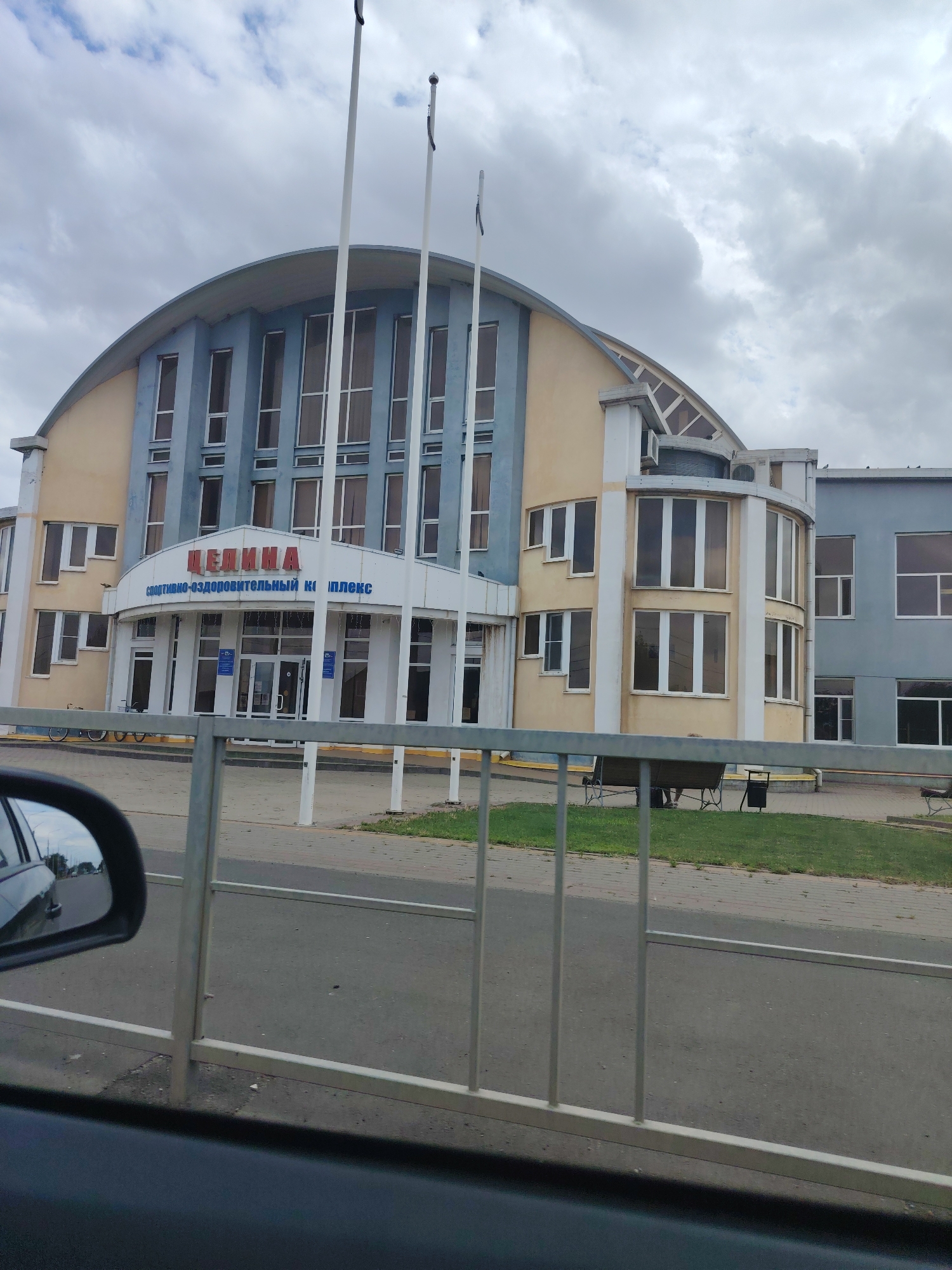 The image shows the exterior of the "Tselina" Sports and Health Complex, located in the city of Tselina, Russia. The building features a prominent arched roof and a facade of light blue and beige colors. Large windows are visible on multiple levels. The entrance area is sheltered by a white canopy. Blue signs with text in Cyrillic are affixed to the building. Three flagpoles stand in front of the structure. To the left, a car's side mirror is visible, reflecting the road and sky. A metal fence runs across the foreground. In the background, there is a grassy area with benches and a trash receptacle. The sky is overcast with clouds.