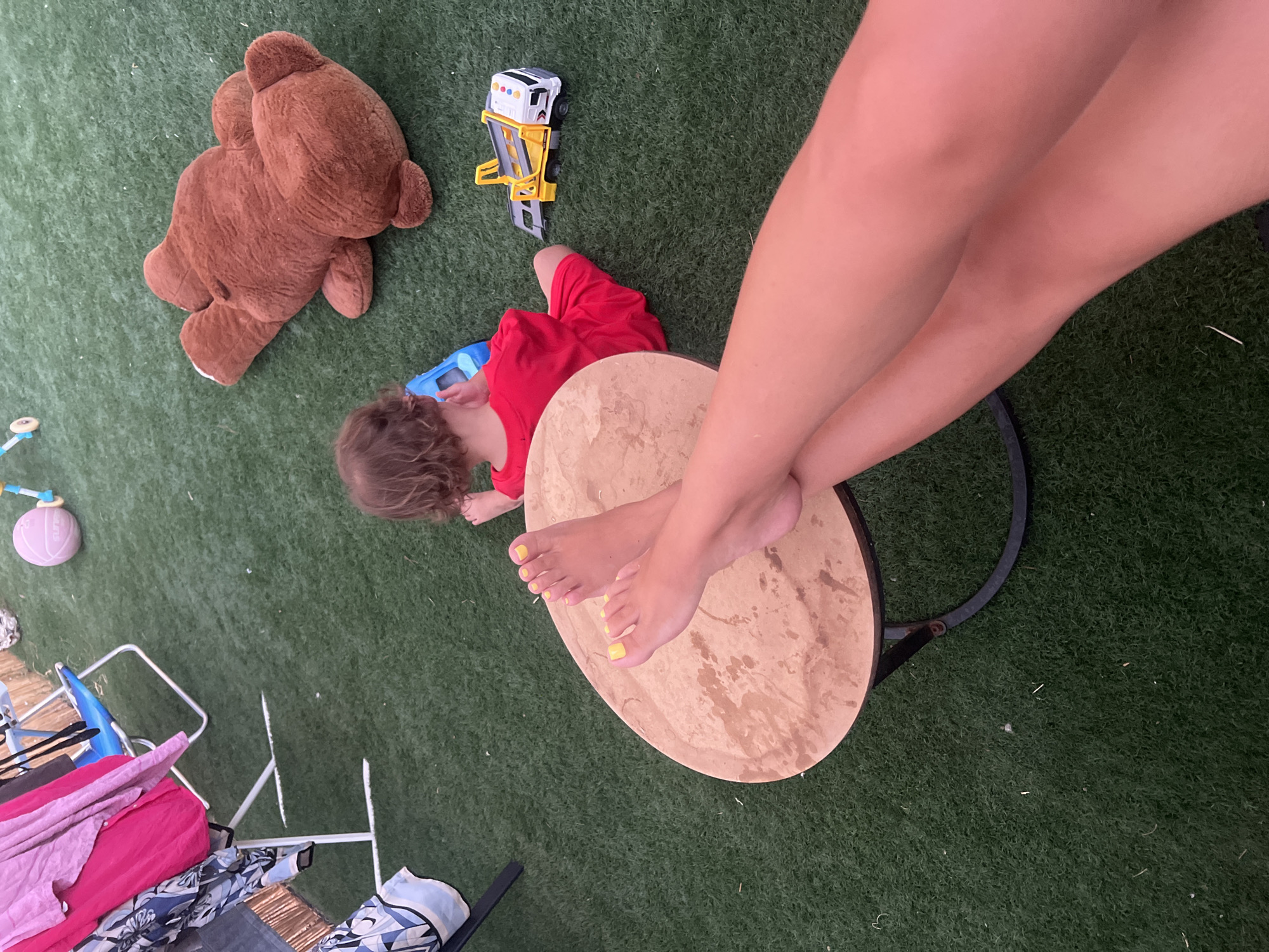 The image captures an outdoor scene on artificial turf, likely in Kiryat Yam, Israel, with a child and a large stuffed bear as the main subjects. The child, seen from behind, is wearing a red outfit and appears to be playing with a blue toy. A brown stuffed bear is lying on the ground next to the child. In the foreground, a person's legs and feet are visible, with bright yellow nail polish on their toes, resting on a circular wooden table. Various items, including clothes on a drying rack and a toy truck, are scattered around the area, suggesting a playful or domestic setting. The overall atmosphere seems casual and relaxed.