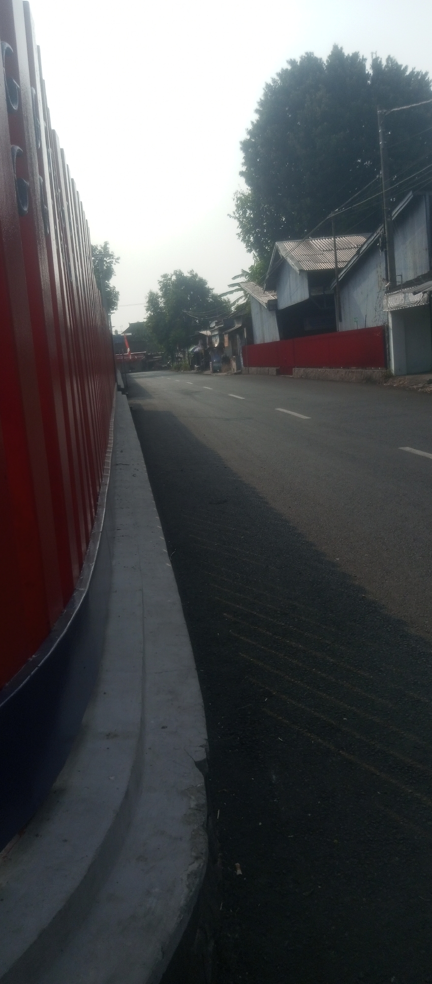 The image shows a street scene in Kediri City, Indonesia. On the left, a tall, corrugated red wall runs along the side of the road. The wall has vertical ridges and appears to be made of metal. A grey concrete curb separates the wall from the asphalt road. The road itself is dark grey and has faint white lane markings.
In the background, there are buildings with corrugated roofs, typical of residential or commercial structures in the area. Trees are visible behind the buildings, and a large, dense tree with dark green foliage dominates the upper right portion of the frame. A utility pole with wires is also present near the large tree. The sky is overcast and bright, suggesting it is daytime, possibly early morning or late afternoon due to the lighting. There are no discernible people or vehicles actively engaged in any activity in the visible parts of the scene. The overall impression is a quiet street on a hazy day.