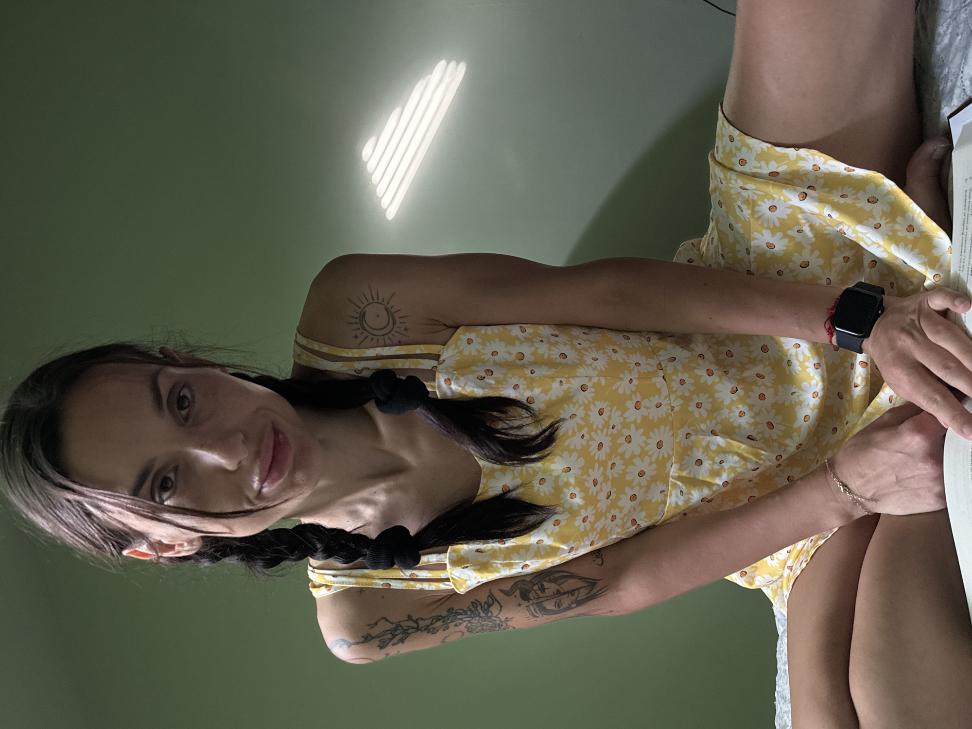 A woman is sitting indoors, wearing a yellow dress with a white daisy pattern. Her hair is styled in two braids tied with black bands. Tattoos are visible on both arms. She is wearing a watch on her left wrist and a bracelet on her right wrist, and she appears to be reading a book. The wall behind her is a muted green color with a pattern of light coming through blinds. The location in the city of Pleven, Bulgaria is not discernible from the image.