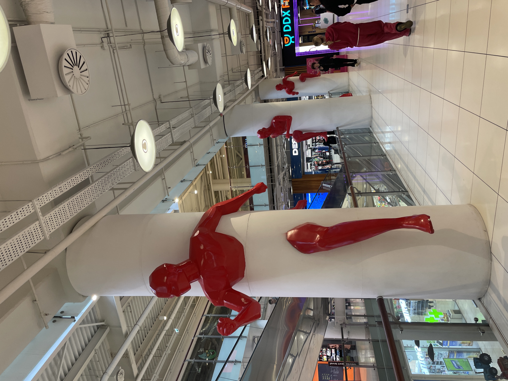 The image shows an interior space, likely a shopping mall, in Khimki, Russia. Dominating the scene are large, white cylindrical columns decorated with bright red, fragmented figures of a climbing man. The columns appear to be structural elements within the building.
Several people are visible in the background, some walking and others standing. The flooring is composed of light-colored tiles. Above, the ceiling is a complex network of pipes, lights, and ventilation systems. In the background, there are storefronts with illuminated signs, including one that reads "DDX". Escalators are also visible, further indicating a multi-level shopping environment.