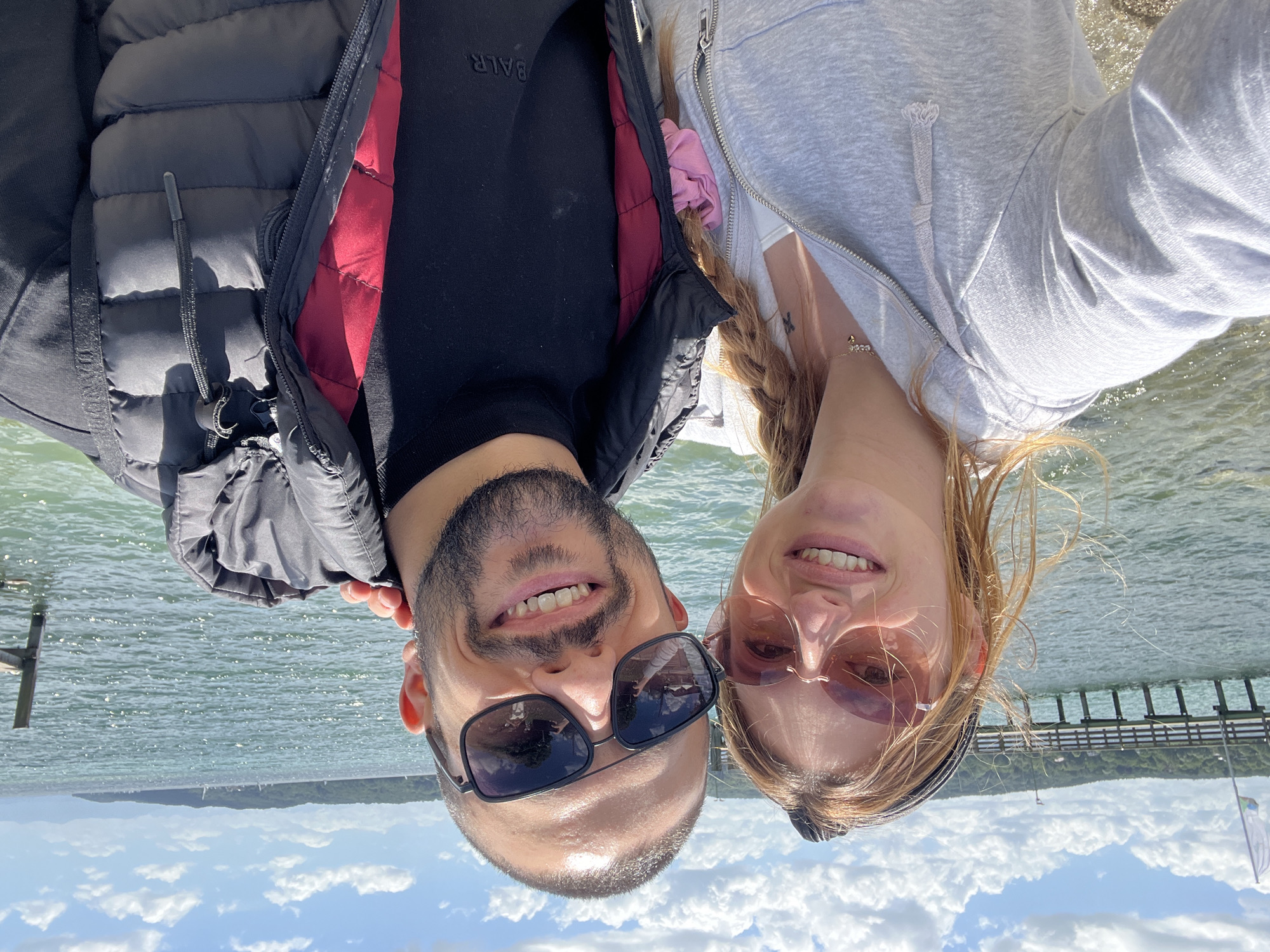 A selfie is taken by a man and a woman on a sunny day by a body of water. The man is on the right, wearing sunglasses, a black puffer jacket with a red lining, and a black t-shirt. The woman is on the left, wearing pink rimless sunglasses, a grey zip-up hoodie, and a white top. She has blonde hair tied back with a black headband, and a pink scrunchie is visible on her hoodie. Both are smiling. Behind them, the water is choppy, and a wooden pier extends into the lake. In the background, a tree-lined shore and distant hills are visible under a blue sky with white clouds. The scene appears to be on the Starnberger See in Germany.