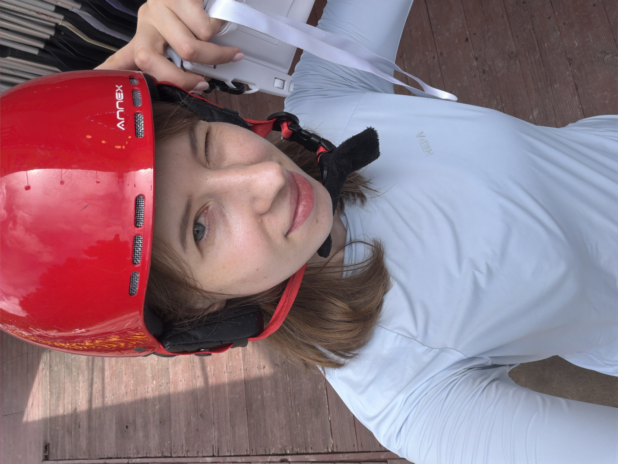 A young woman, wearing a red helmet with "ANNEX" written on the side, and a light blue long-sleeved shirt, is taking a selfie outdoors. She is winking at the camera and holding a white object with straps. The background appears to be a wooden deck, possibly a porch or boardwalk. The lighting suggests it is daytime. There are hangers with clothing visible in the upper right corner of the image. There are no other people visible in the frame. The location is not specified in the image.