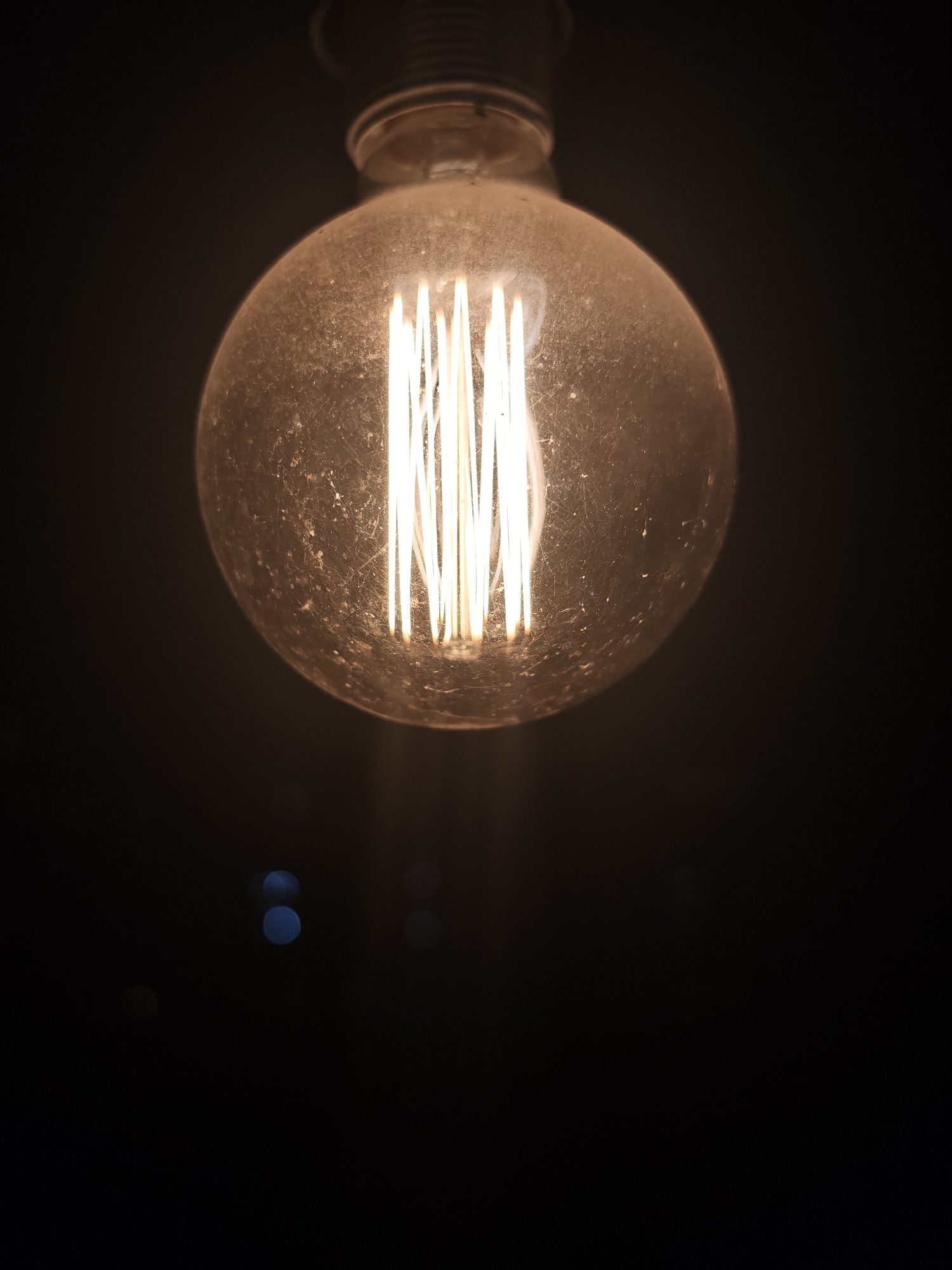 The image shows a close-up of a vintage-style lightbulb with multiple vertical filaments, emitting a warm yellow light. The bulb itself appears slightly dusty or smudged, adding to its aged aesthetic. The background is dark, almost completely black, with a few out-of-focus blue and gray bokeh lights visible in the lower portion of the image. The bulb is centered in the frame, and the composition directs focus entirely on the light and its immediate surroundings. There is no evidence that this image was taken in Moscow, Russia.