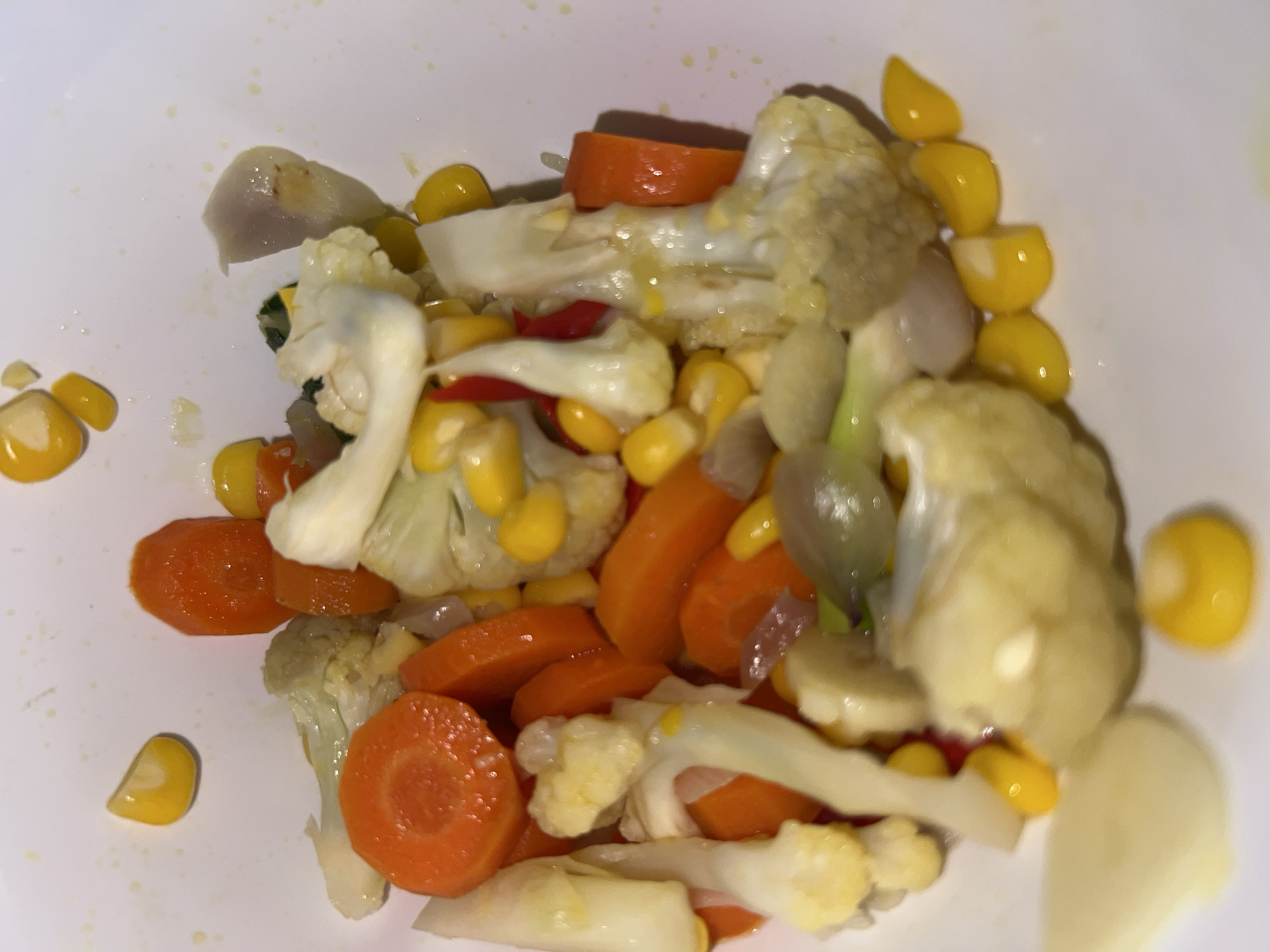 This is a close-up, top-down shot of a white bowl filled with cooked mixed vegetables. The vegetables include bright orange carrot slices, white cauliflower florets, kernels of yellow corn, and pieces of thinly sliced red onion. Some of the corn kernels are broken. The vegetables appear to be lightly coated in a clear liquid, possibly water or oil, with a few small yellow droplets scattered on the white surface of the bowl. The background is a plain white. There are no people or discernible environmental features related to Klapanunggal, Indonesia, present in the image.