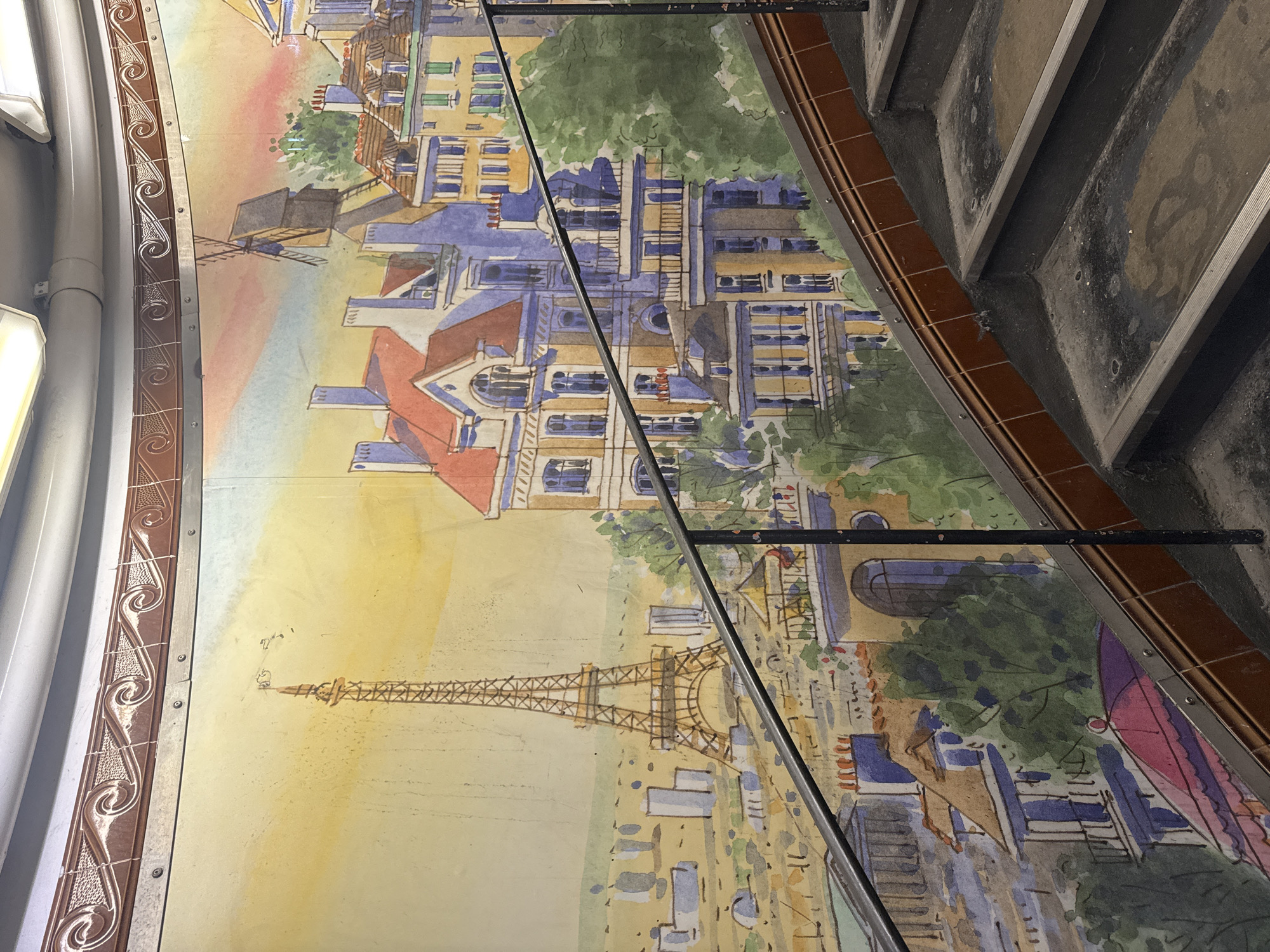 The image shows a mural of Paris, France, featuring the Eiffel Tower and a windmill, likely the Moulin Rouge. The mural is painted on a curved surface, possibly a wall or ceiling, and is bordered by decorative trim. In the foreground, there are elements of architecture, including a staircase and a metal support beam.