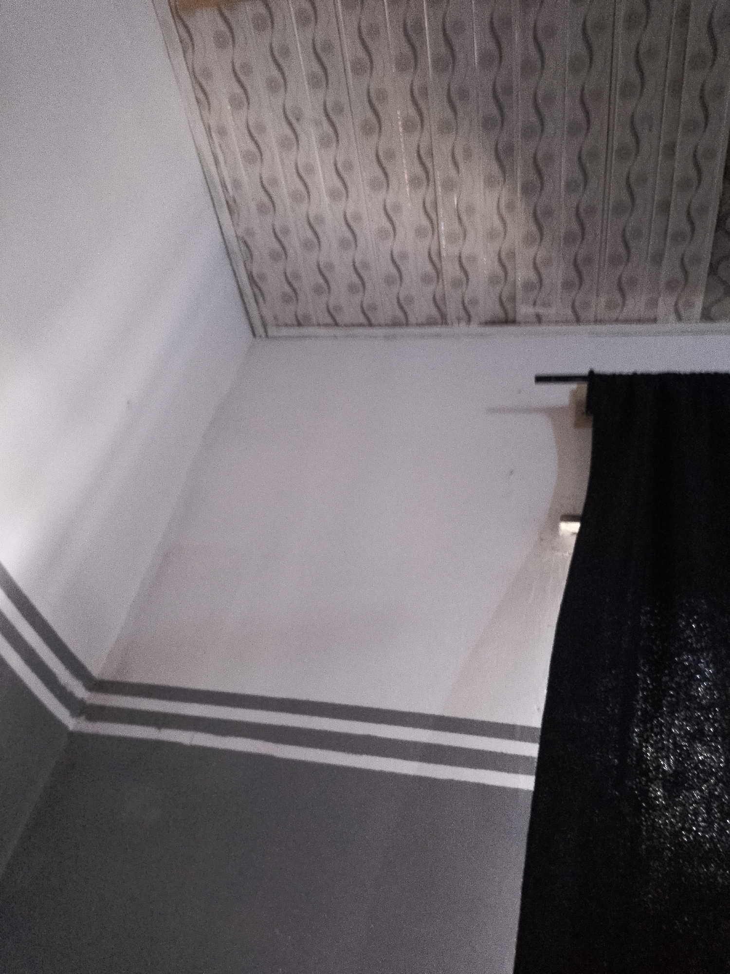 The image shows an interior scene with a white ceiling that has a repeating wavy pattern. The walls are painted in two tones of gray, with two horizontal white stripes separating the lighter upper section from the darker lower section. On the right side of the image, a black curtain hangs from a dark rod. The lighting in the room is soft, casting shadows on the walls and ceiling. There are no people or any discernable objects other than the architectural features and the curtain. The scene appears to be a bedroom or a living area.
