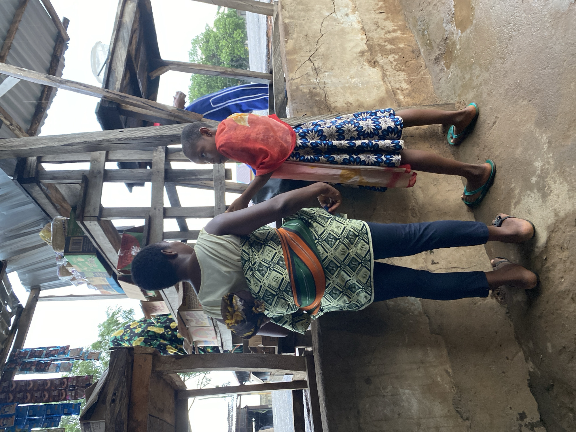 A woman, from the back, is carrying a baby wrapped in a patterned cloth on her back. She is wearing a light-colored sleeveless top and dark pants, with sandals. A young girl, facing away from the camera but looking to her left, stands beside her. The girl is wearing a blue and white floral dress and a red and pink scarf draped over her shoulders. They are standing in an outdoor market setting with wooden structures, shelves stocked with goods, and a rough concrete or dirt ground. The background shows trees and other buildings.