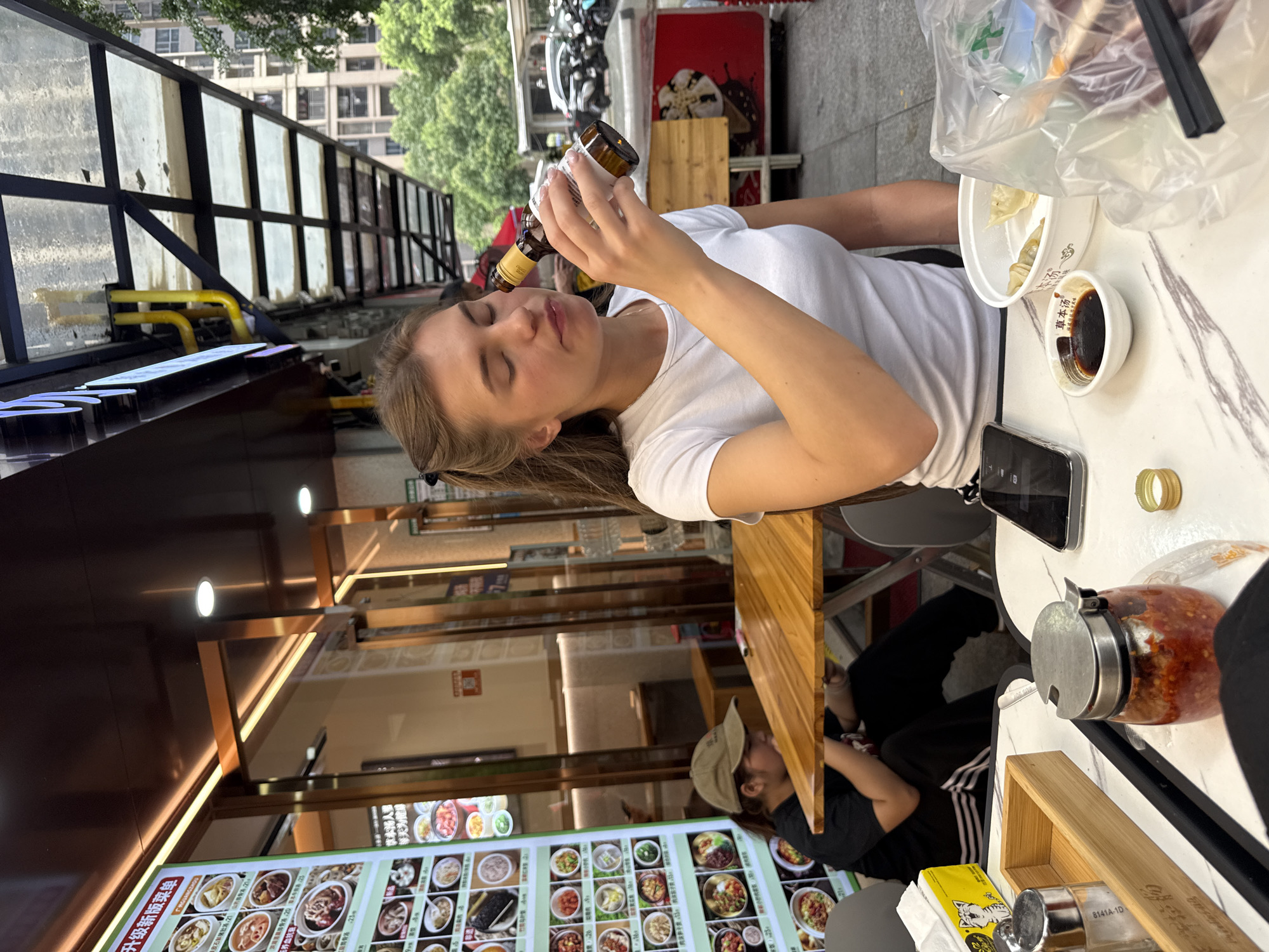 In Haizhu District, China, a young Caucasian woman with blonde hair pulled into a ponytail, wearing a white t-shirt, is holding a small brown bottle to her lips as if to drink from it. Her eyes are closed, and her lips are slightly parted. She is sitting at a marble-topped table outdoors, on a sidewalk in front of a restaurant. To her left, another person, possibly a woman, wearing a beige cap and a black t-shirt, is seated at the same table, facing away from the camera and looking down. The table is laden with food and condiments, including a jar of chili paste, a bottle of soy sauce, a bowl of dumplings, and a cell phone. In the background, a menu is displayed on a wall inside the restaurant, showing various food items. A covered awning extends over the seating area, with utility pipes visible. Beyond the awning, trees and apartment buildings can be seen, along with parked motorcycles. The scene appears to be a casual dining experience on a street in China.