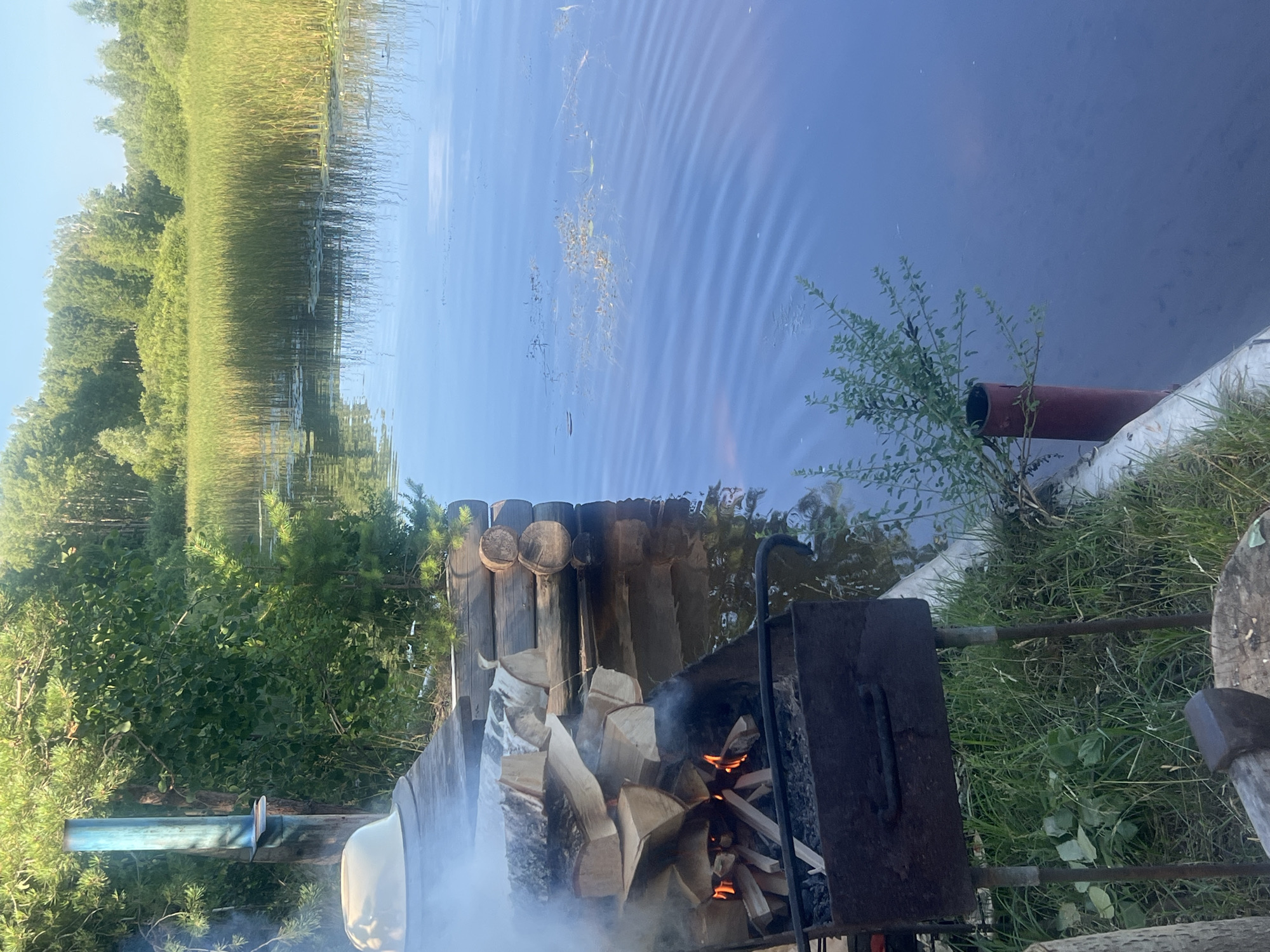 The image depicts a serene lakeside scene in Chaykovsky, Russia, on a clear day. In the foreground, a makeshift grill with burning wood and logs is situated on a wooden dock, with smoke rising from the fire. The dock is constructed of rough-hewn logs and planks, and next to it, a pile of firewood is neatly stacked. Beyond the dock, a calm body of water stretches out, reflecting the blue sky and the lush green trees lining the distant shore. The water is interspersed with patches of reeds and aquatic plants, creating a natural, untouched ambiance. The overall impression is one of tranquility and rustic simplicity, suggesting a peaceful day spent outdoors by the water.