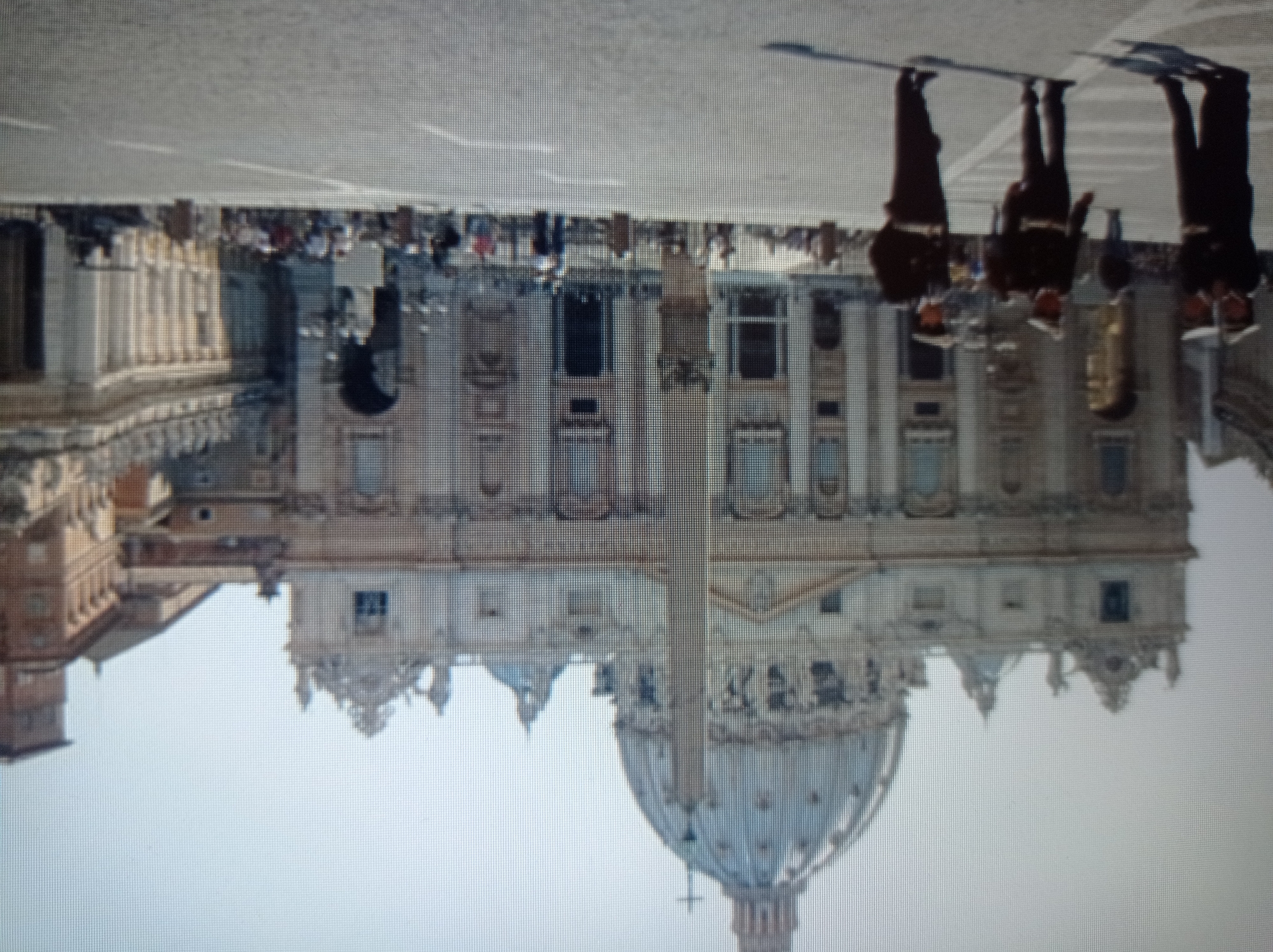 The image depicts a view of St. Peter's Basilica in Vatican City, Rome, Italy, not Mukachevo, Ukraine. The foreground shows three individuals in dark uniforms with white belts and hats, appearing to be guards or police officers, standing in a large, open square paved with cobblestones. Behind them, the grand facade of St. Peter's Basilica is visible, dominated by its dome and an obelisk in front. The architecture is classical, with columns, arches, and intricate detailing. The background shows more of the basilica's structure, including smaller domes and a clock tower, as well as other buildings. Scattered throughout the square are other figures, appearing to be tourists or pedestrians, and some benches or planters. The lighting suggests it is daytime, with the sun casting shadows. The overall scene conveys a sense of grandeur and public space.