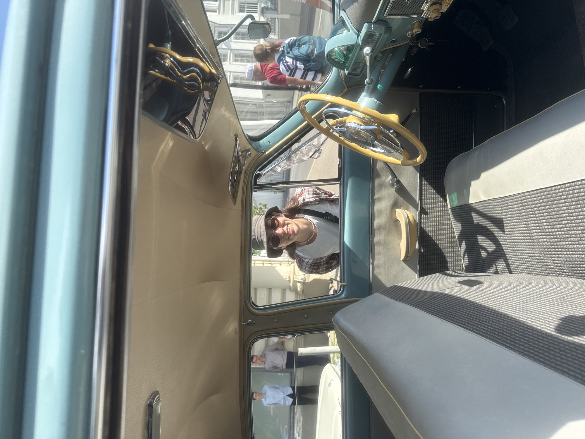 The image shows the interior of a light blue vintage car, possibly a convertible, with beige interior trim. The car has a light-colored steering wheel with chrome accents. Through the car windows, a woman wearing sunglasses and a hat is visible, and she appears to be looking into the car. Reflections in the windows show people standing outside near a building, suggesting an urban environment. The seating is light grey with patterned inserts. The car's dashboard is visible, with various knobs and controls. The location is likely Kazan, Russia, based on visual clues that may be associated with the city.