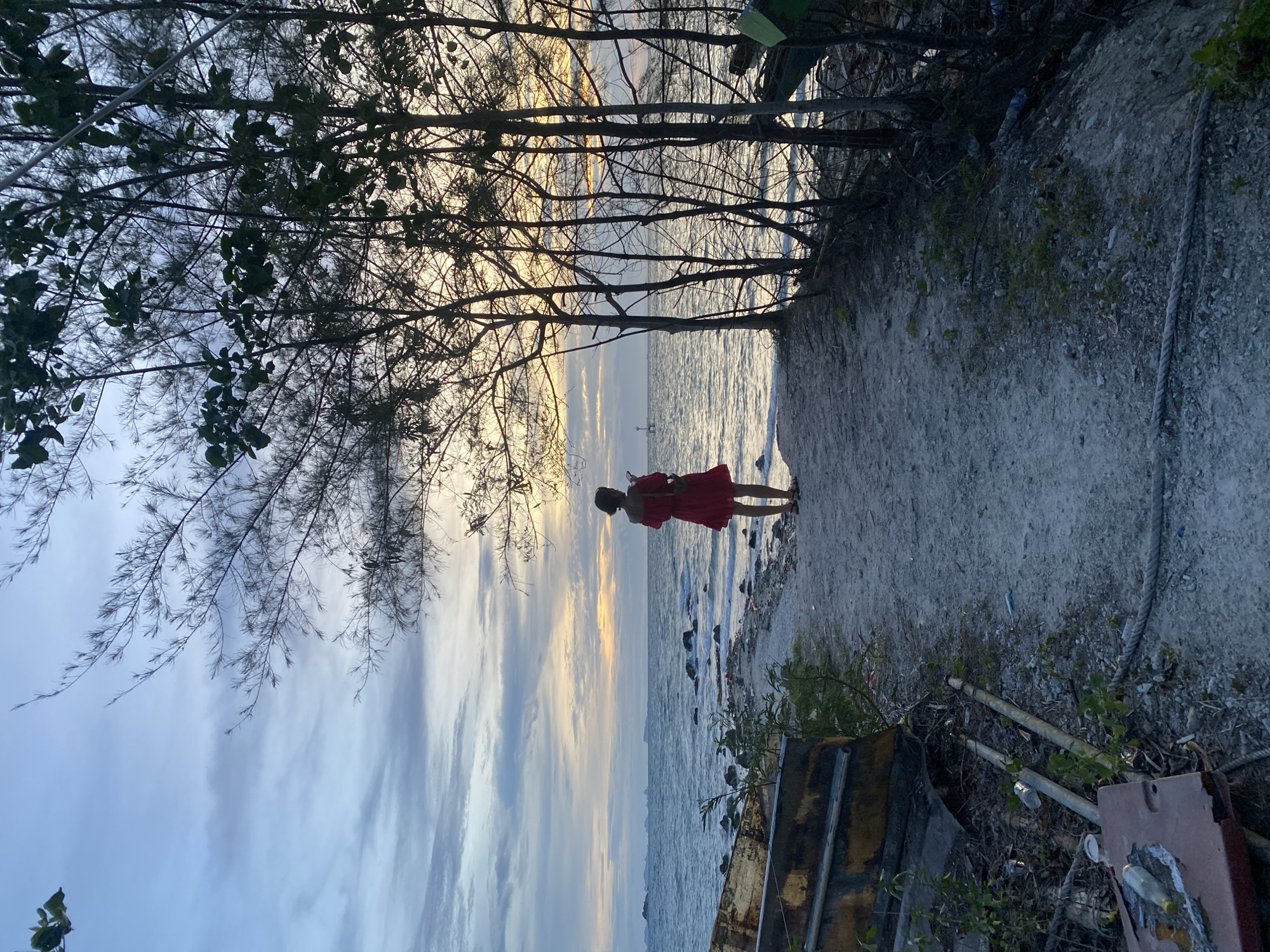 A person in a red dress stands on a sandy path looking out at the ocean. The person is facing away from the camera, holding a phone in their right hand. Behind the person, the ocean meets the horizon under a cloudy sky with streaks of orange and yellow from the setting sun. Several tall, thin trees with sparse foliage line the right side of the path. In the foreground, to the left of the path, lies a weathered, yellow boat hull. The ground is a mixture of sand, pebbles, and scattered debris, including plastic bottles, cans, and rope. The overall impression is a serene, natural environment during twilight, with a hint of disarray from the debris.