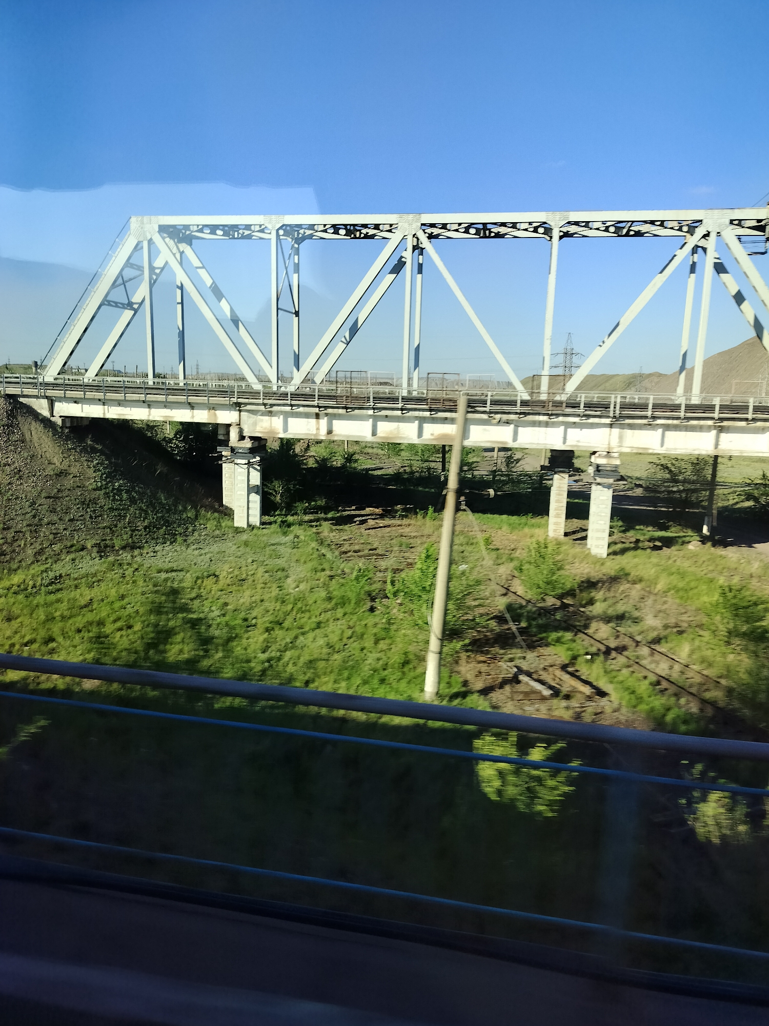 A view of a railway bridge from a moving vehicle. The bridge is a light gray truss structure with a roadbed running between the supports. The bridge spans a grassy area with scattered trees and bushes. A wooden utility pole stands near the center of the image. In the background, utility poles and a small, rocky hill are visible. The sky is clear blue. In the foreground, a blue guardrail indicates the viewer is likely on a train or bus. The location is Unknown, Unknown.