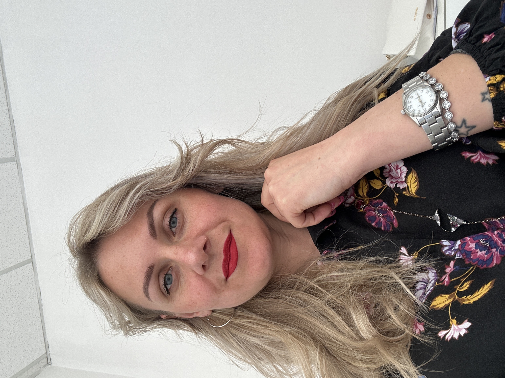 An adult woman with blonde hair and blue eyes is depicted in a close-up, rotated image. She looks directly at the viewer with a pleasant, slight smile, and red lipstick. Her left hand is raised near her temple, forming a loose fist.
She is wearing a black top or dress featuring a vibrant floral pattern of purple, pink, and yellow flowers. On her left wrist, she sports a silver-toned watch with a round face and a metal link band, accented with small clear stones around the bezel. Adjacent to the watch is a silver-toned bracelet adorned with multiple clear, circular stones. A subtle star-shaped tattoo is partially visible on her inner left forearm. Silver-toned hoop earrings are visible, and a thin silver-toned necklace with a small, gem-decorated pendant (possibly a butterfly) hangs near her shoulder.
The background is predominantly a plain, light-colored surface, likely an indoor wall or ceiling, with what appears to be part of a ceiling grid with square tiles visible in the upper left corner of the rotated image. The lighting is bright and even, suggesting an indoor setting during daytime. The woman appears to be posing for the photograph. No discernible text is clearly visible in the image. The specified location context is Venafro, Italy, but no visual cues in the image confirm this.