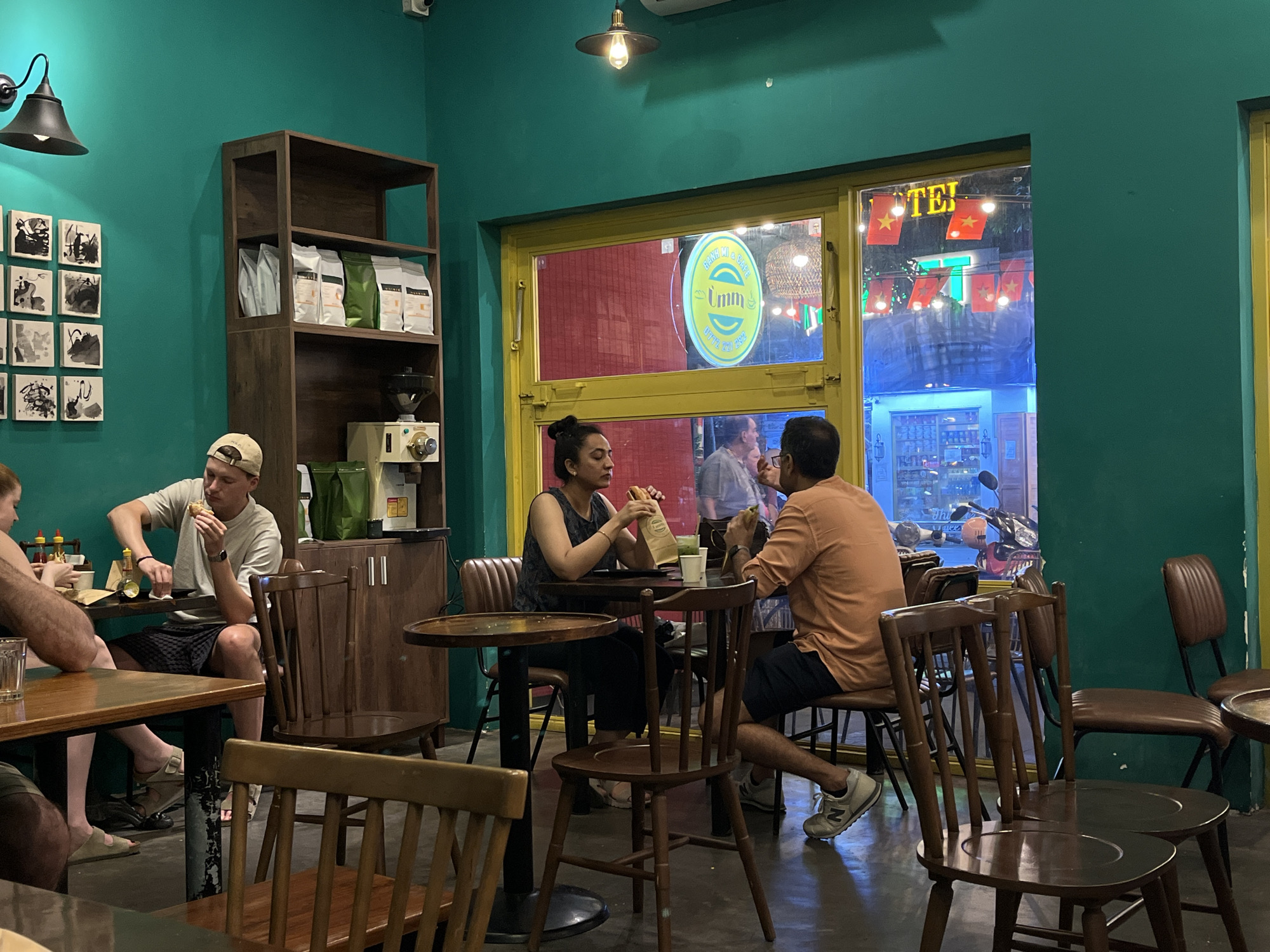 Captured indoors, a café in Đà Nẵng, Vietnam is active. A window prominently displays "Umm" signage and red Vietnamese flags. Patrons are seated at various wooden tables and chairs, some eating. A man with a beige baseball cap eats while seated near a shelf stocked with coffee and an espresso machine. Another pair is seated in the middle, eating. Other customers are present, partially visible. The café has turquoise walls.