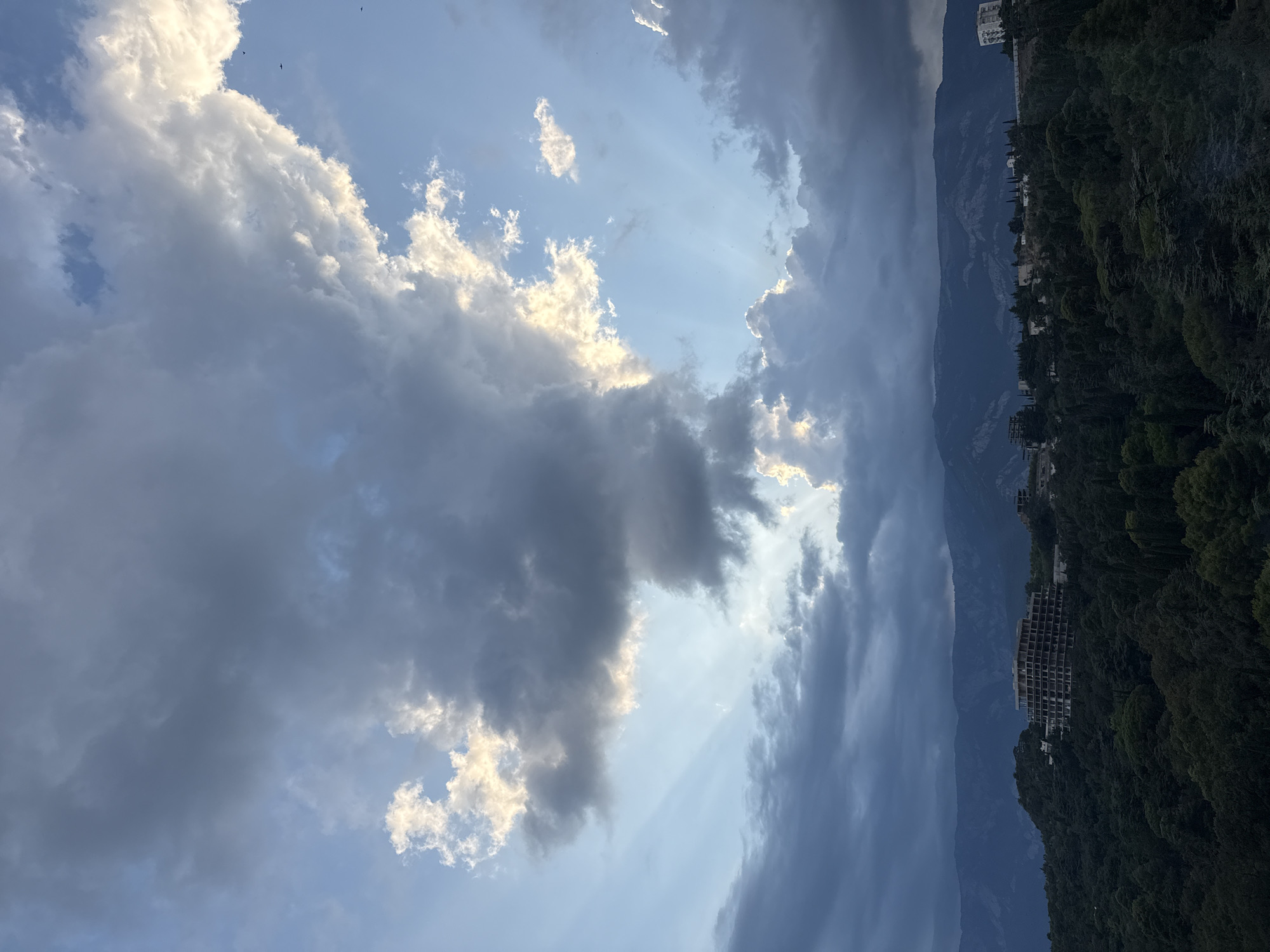 The image captures a dramatic sky over a city or town nestled amongst hills and mountains, likely in Yalta, Ukraine. A vast expanse of clouds dominates the upper portion of the frame. Some clouds are dark and heavy, suggesting an impending storm or the remnants of one, while others are illuminated by sunlight, creating a striking contrast. Rays of light pierce through the clouds in places, forming crepuscular rays that fan out towards the horizon.
Below the sky, a densely forested hillside slopes down towards a body of water or a valley. A distinctive, multi-story building under construction, with a visible concrete framework and numerous balconies, stands prominently on the hillside amidst the trees. Further in the distance, more buildings, some appearing to be residential or hotels, are scattered across the landscape, along with more of the lush, dark green vegetation that covers the terrain. The distant mountains form a rugged, dark backdrop against the dynamic sky. The overall impression is one of natural beauty and urban development coexisting, with the sky providing a powerful and atmospheric element to the scene.