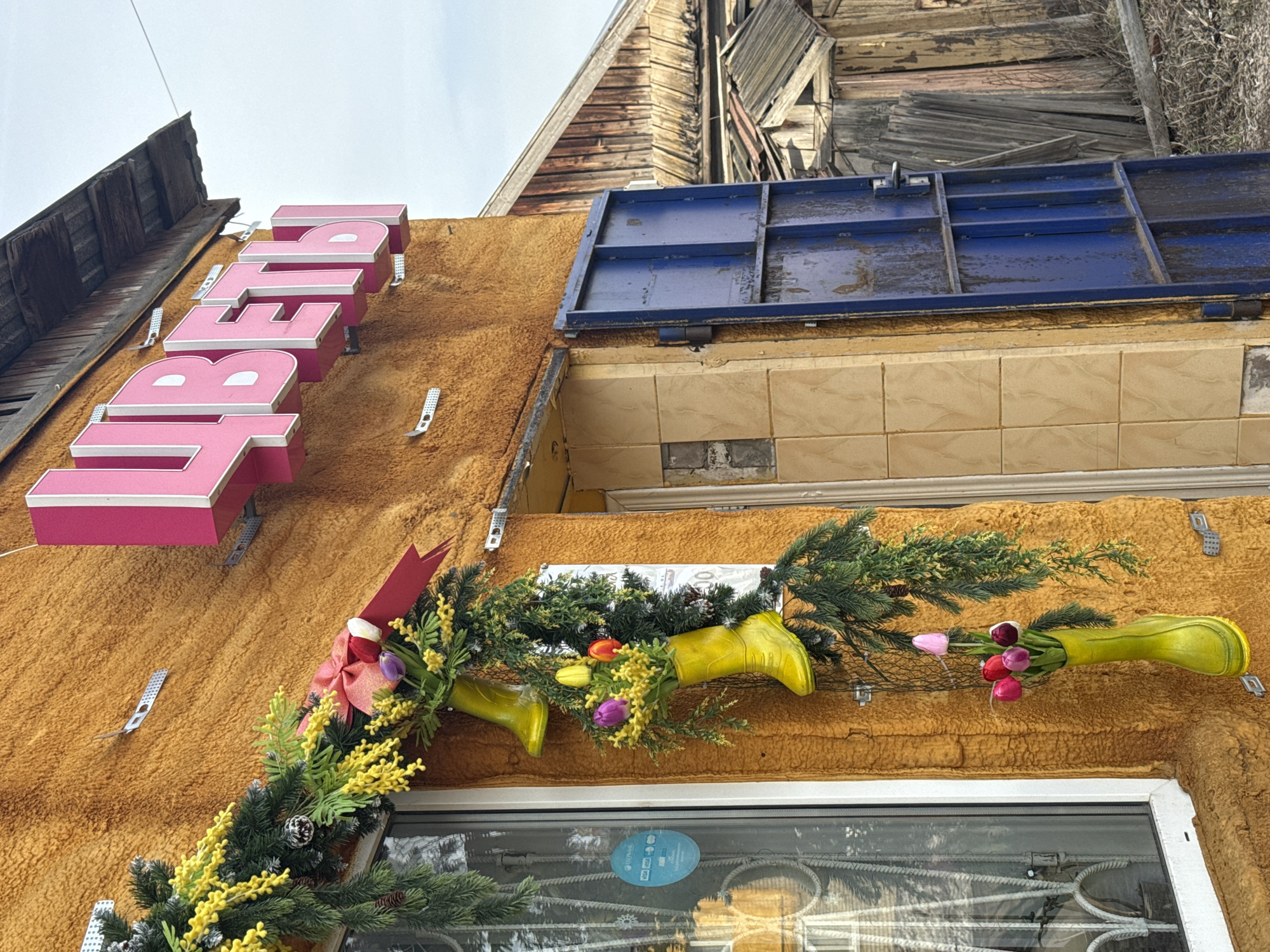 The image depicts the exterior of a building in Фоки, Russia. The building's facade is covered in a textured, orange-brown material, possibly insulation or siding. A pink sign reading "Бедр" is mounted on the wall. The window frame is white, and it is partially covered with a garland and decorations with yellow boots filled with flowers. Above the window, the building is tiled and blue metal storage is mounted at the top. The building has two levels, with weathered wood. The sky is overcast.