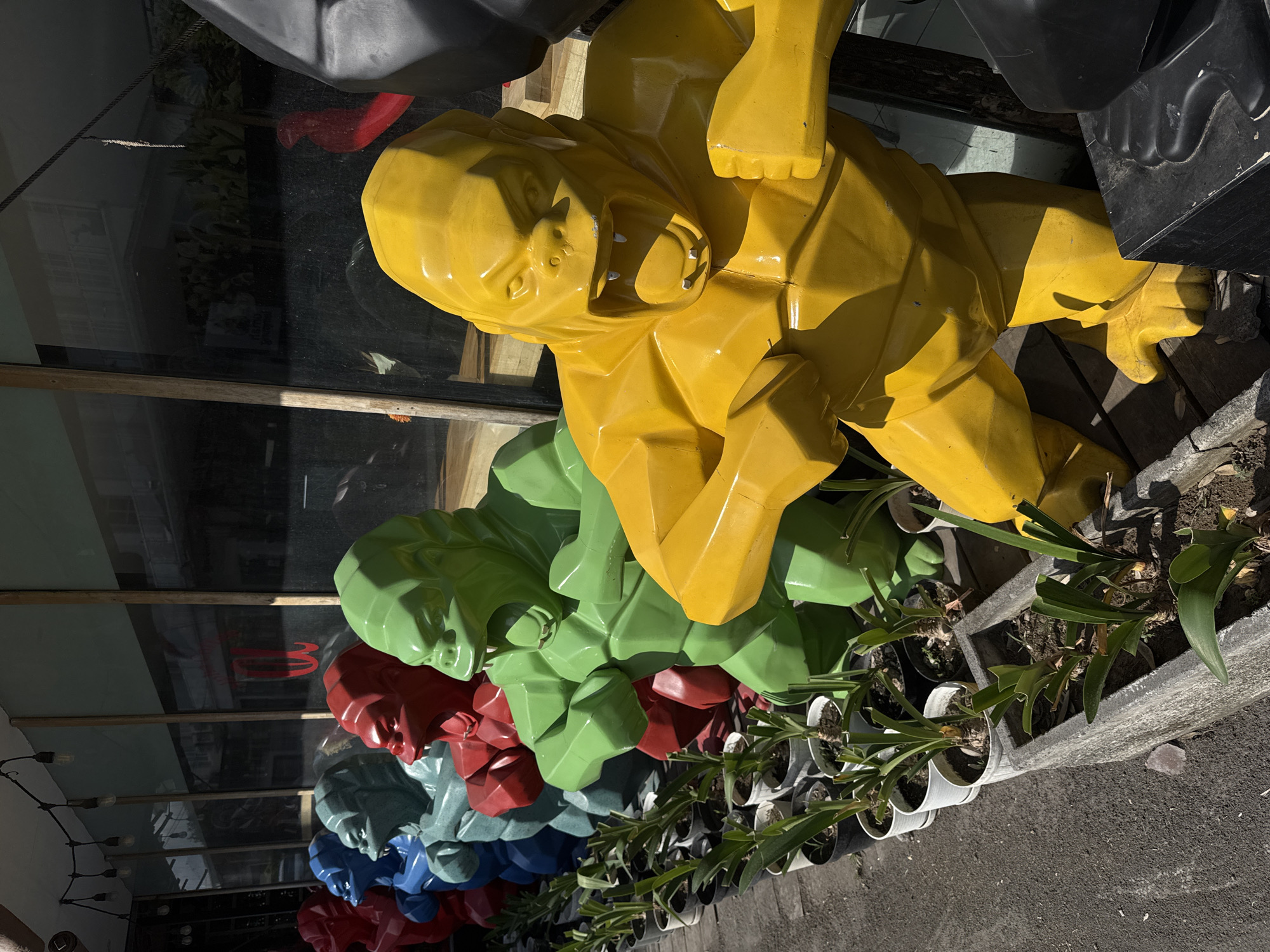In North Kuta, Indonesia, a line of brightly colored, stylized gorilla statues is displayed outdoors. The statues, rendered in red, blue, teal, green, and yellow, are positioned in a row, facing towards the viewer. They have a geometric, faceted design and appear to be made of a glossy material. The yellow statue is most prominent in the foreground, captured in a dynamic pose with its mouth open and fists clenched. Behind the statues, a row of potted plants in white and black planters lines the pavement. A building with large, reflective windows is visible in the background, with some plants and interior details reflected in the glass. Overhead, a string of lights hangs across the scene, casting shadows. The ground is paved with asphalt.