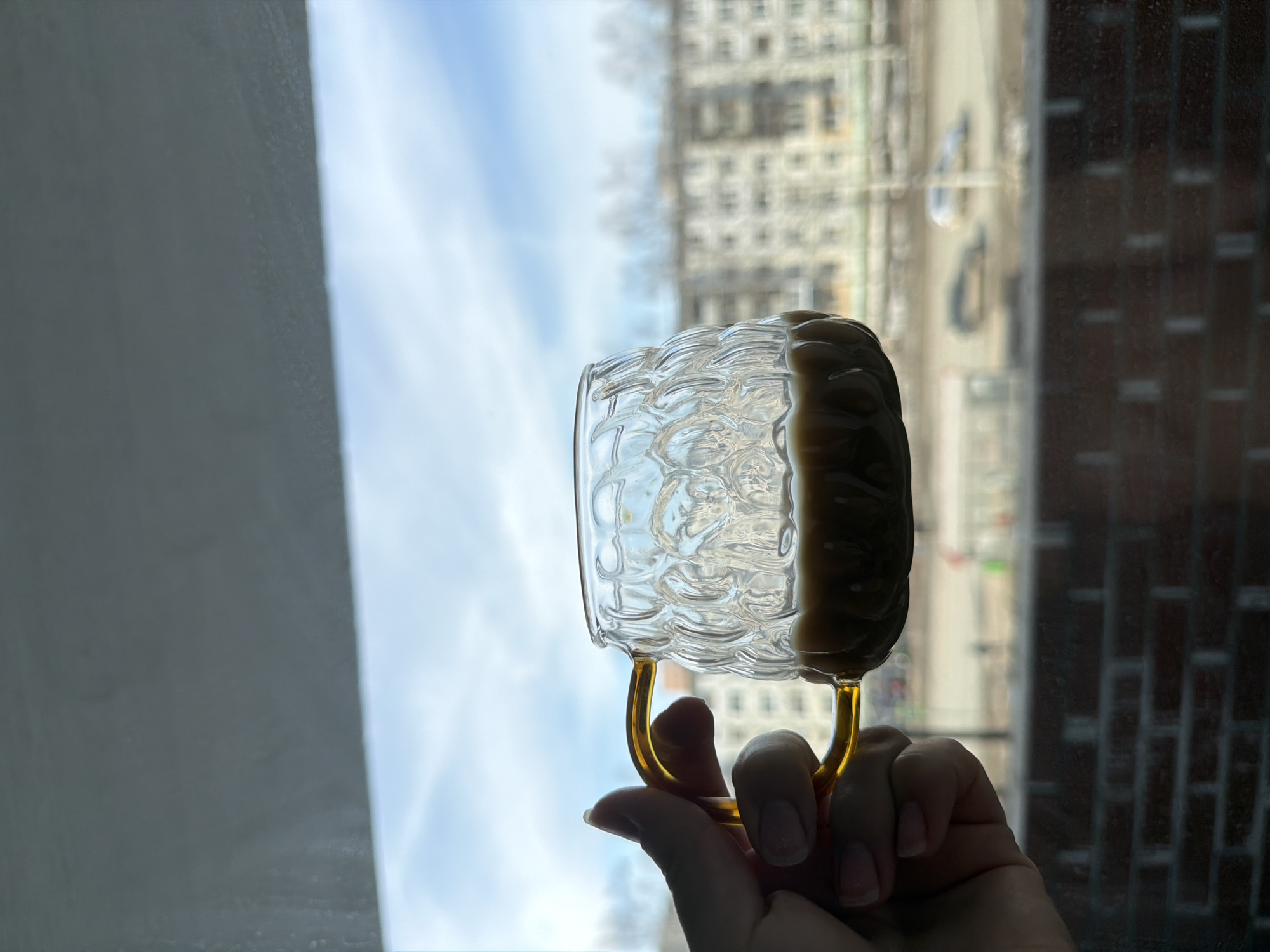 A hand holds a clear glass mug filled with a dark liquid, likely coffee, in front of a window. The mug has a decorative, textured surface and a gold-colored handle. Outside the window, the blurred cityscape of Izhevsk, Russia, is visible, featuring buildings and a cloudy sky. The window frame and a brick wall partially frame the scene.