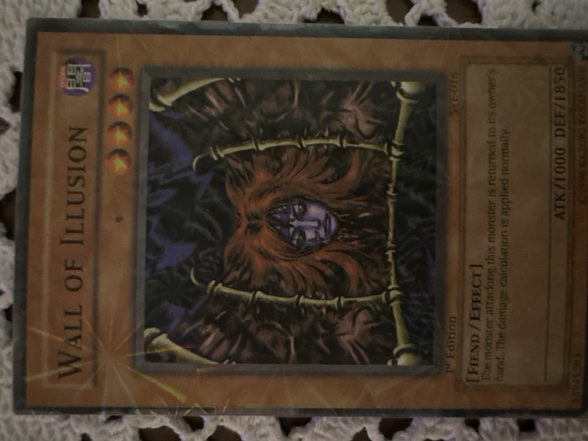 The image shows a Yu-Gi-Oh! card titled "Wall of Illusion." The card is a monster card with the type "Fiend/Effect." The card's attribute icon is visible. The card is titled Wall of Illusion in capitalized letters. The card's Attack and Defense values are 1000 and 1850, respectively. The text "1st Edition" is printed on the card. The card is identified as "SYE-016". The image on the card depicts a monster in a trapped frame.